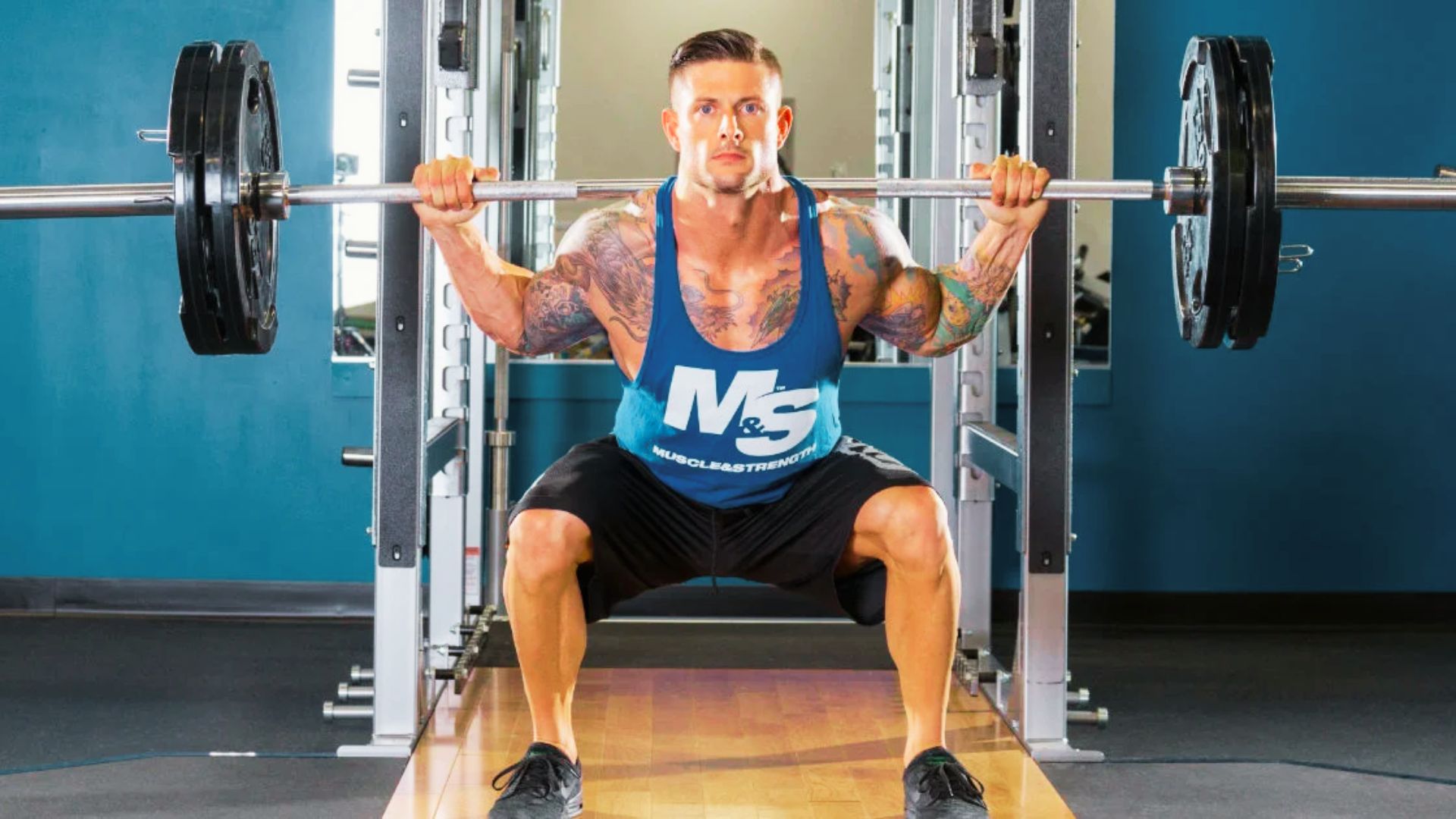 An athletic man with tattoos performs a barbell back squat inside a squat rack.