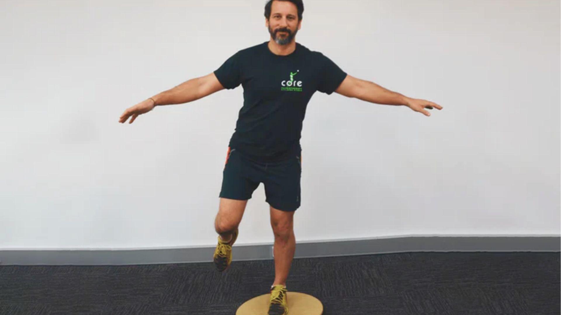 Man with arms extended balances on one leg on a round wooden balance board in a studio.