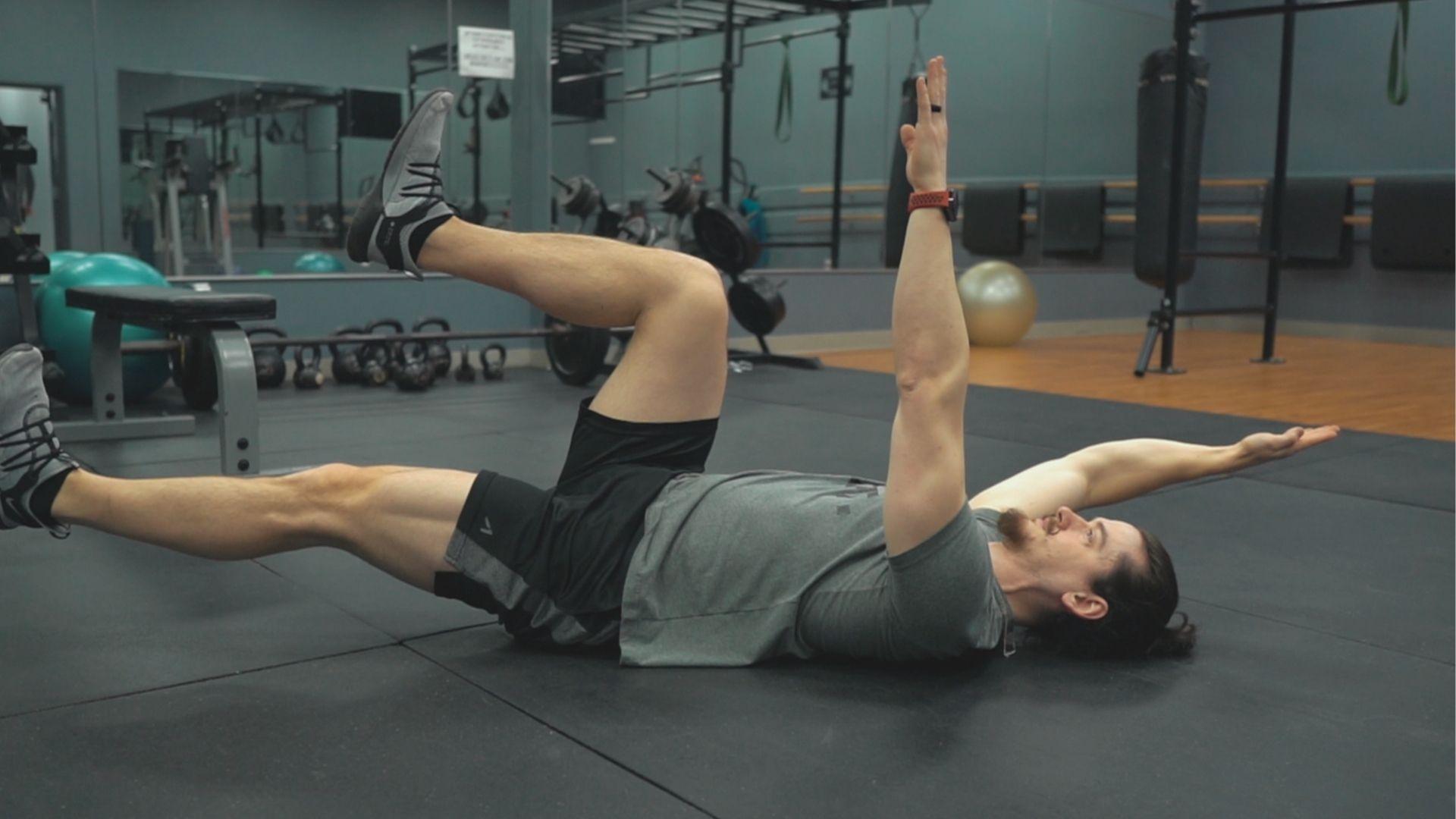 Man in gym performs standard Dead Bug Exercise on black mat with knees bent at 90°.