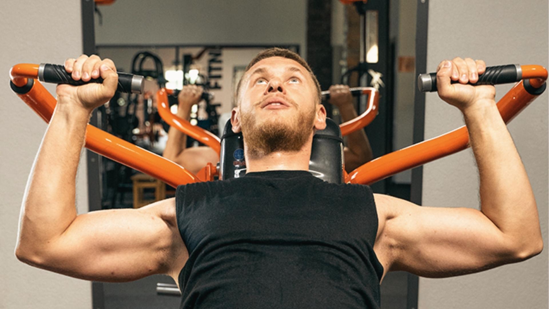 Bearded man in black sleeveless shirt performs machine shoulder press with orange handles, targeting deltoids in commercial gyms.
