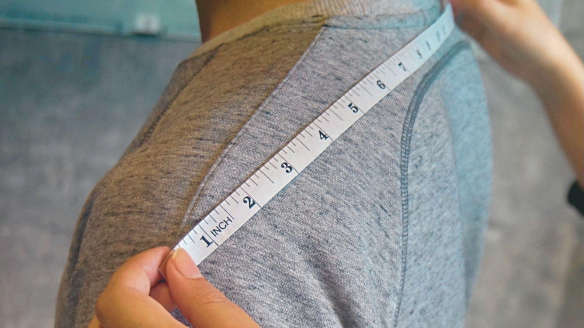 Hands using a white measuring tape to measure shoulder width on a person wearing a gray raglan shirt.