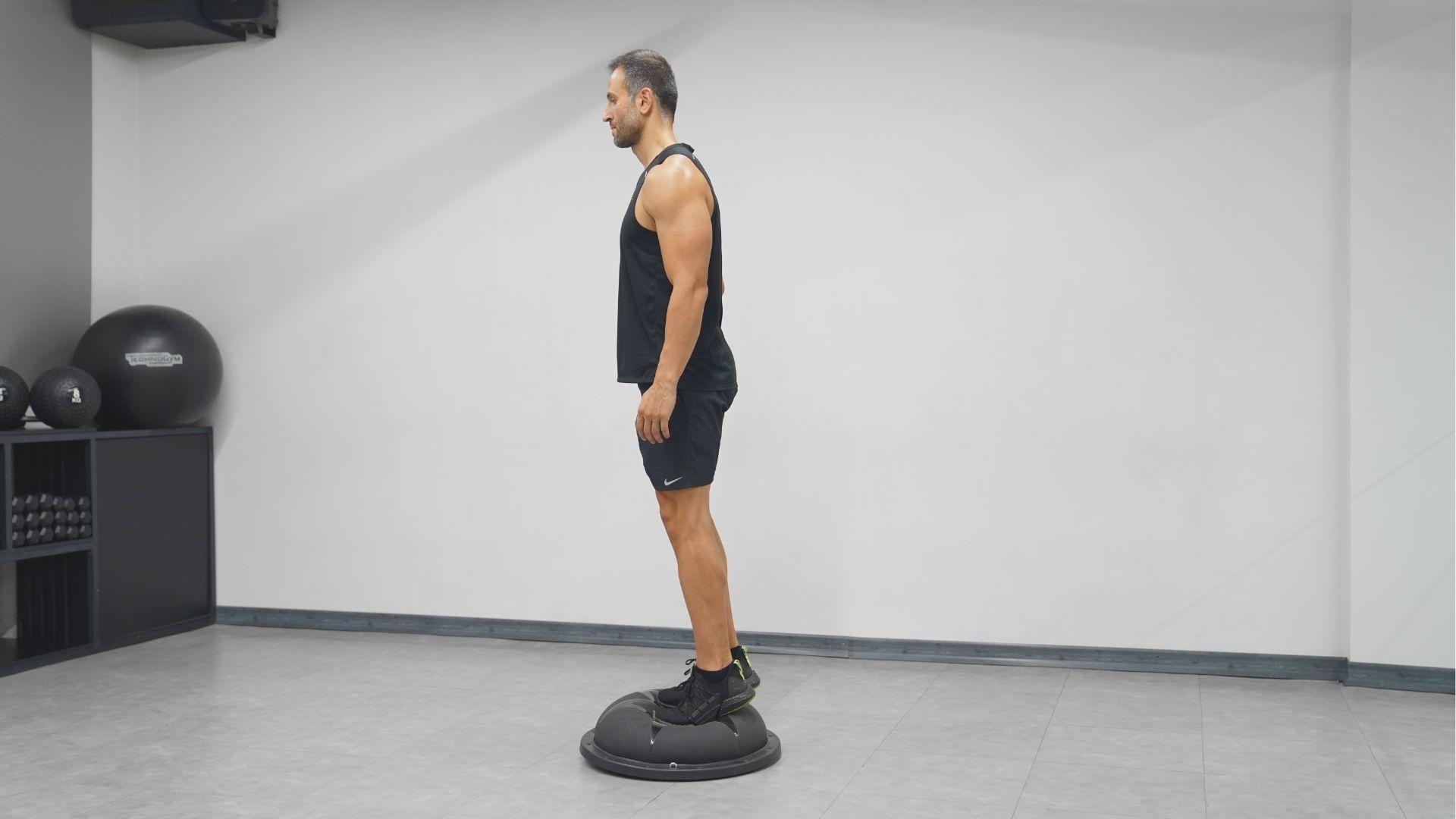 Man in black tank top stands on a BOSU ball with feet together in a gym.