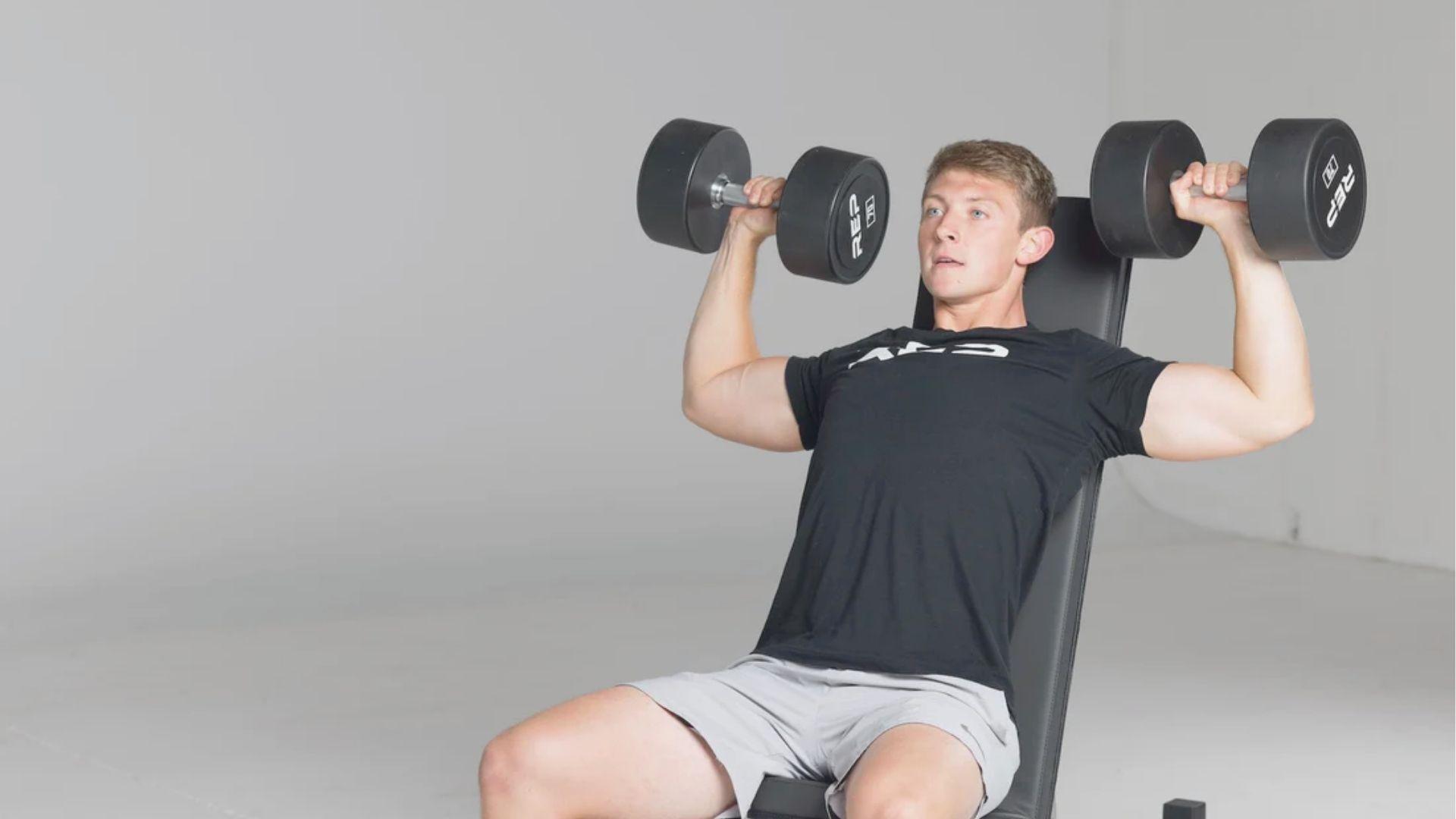 Man in a black t-shirt and gray shorts pressing heavy dumbbells upward while seated on an incline bench.