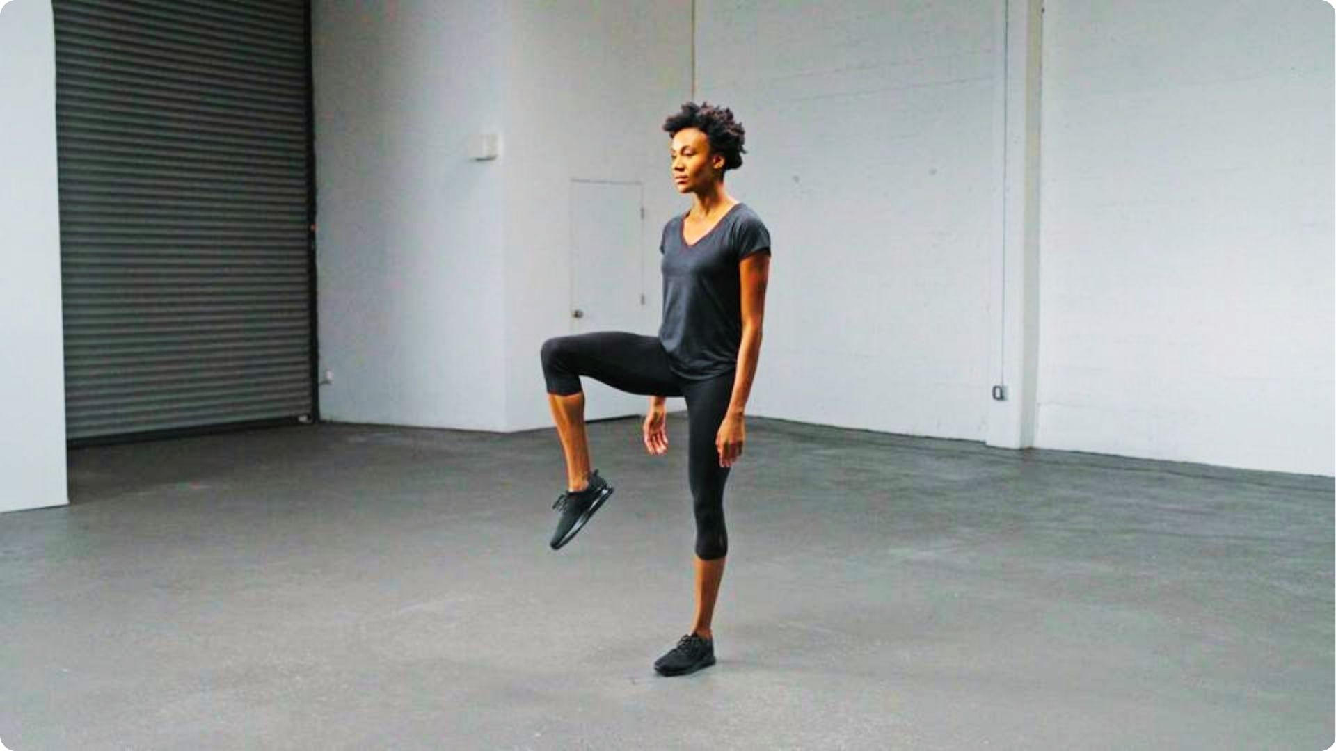 Woman in black workout clothes standing in a studio, lifting one knee up toward her chest.