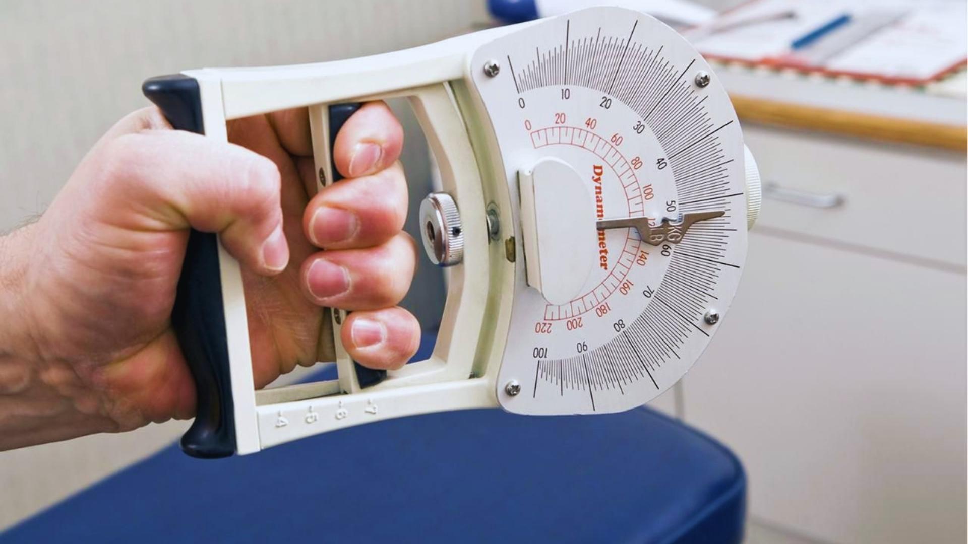 Close-up of a man's hand squeezing a white analog hand dynamometer displaying grip strength in a medical office setting.