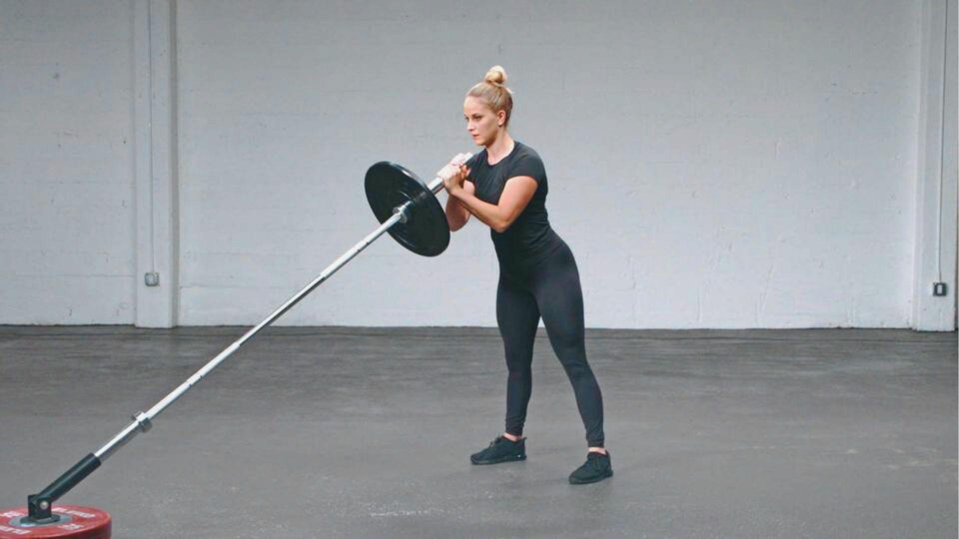 A woman in black leggings and a black tee stands at the top of a landmine squat, barbell held close to her chest.