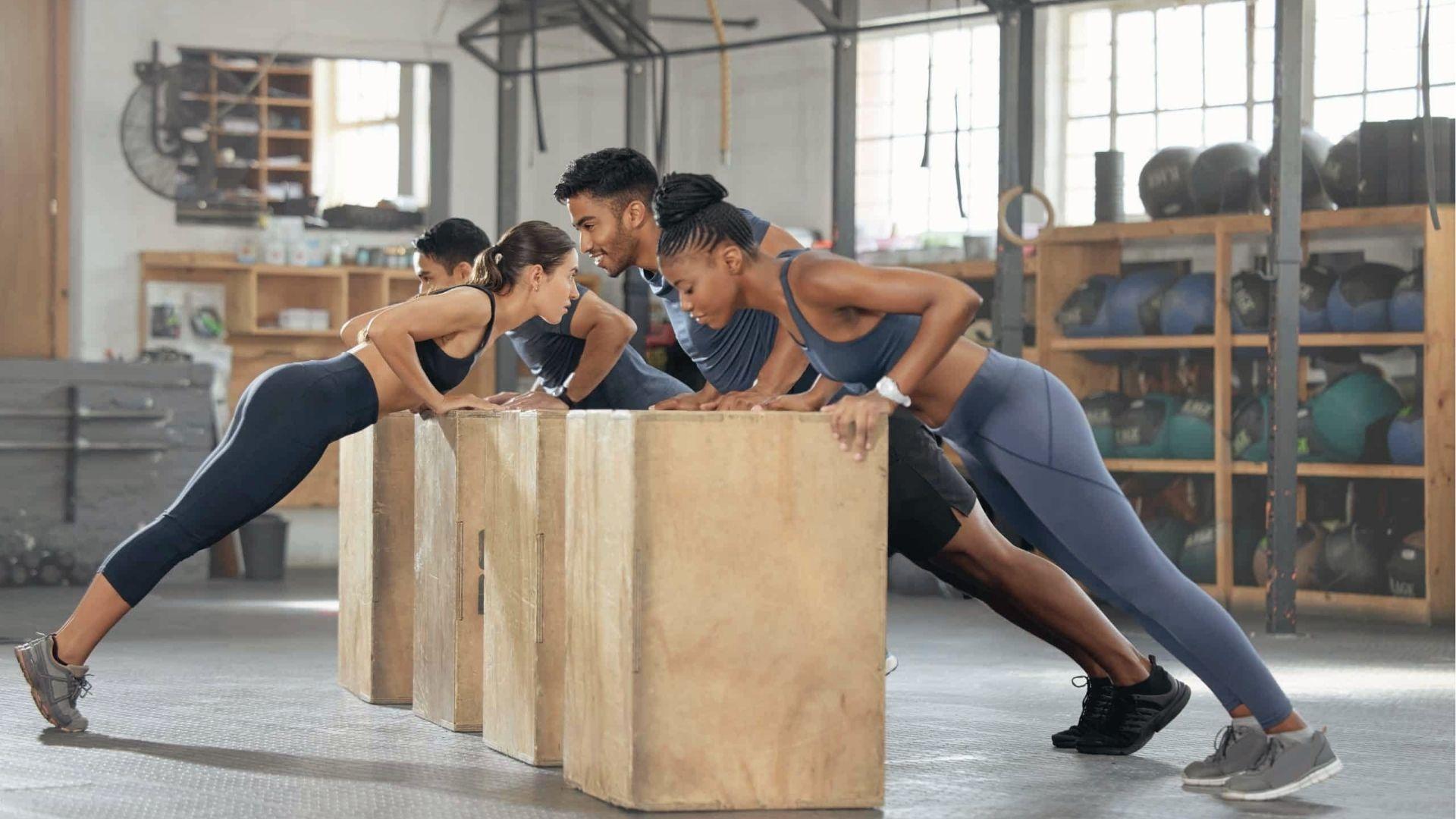 Four people in workout clothes do incline push ups together on wooden boxes inside bright gym