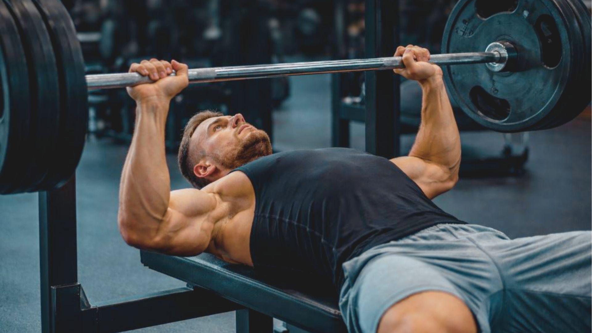 Man in a black tank top lying on a flat bench, pressing a loaded barbell directly above his chest.