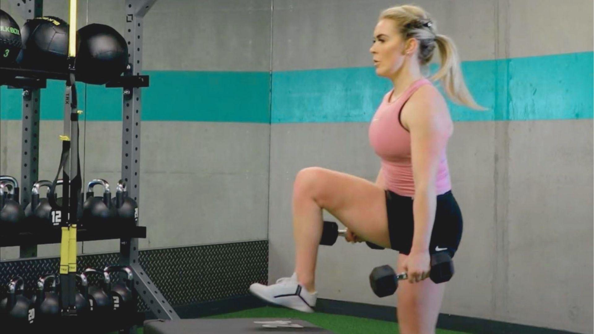 Woman in pink tank top performs dumbbell step-ups on a box in a gym, holding weights at her sides.