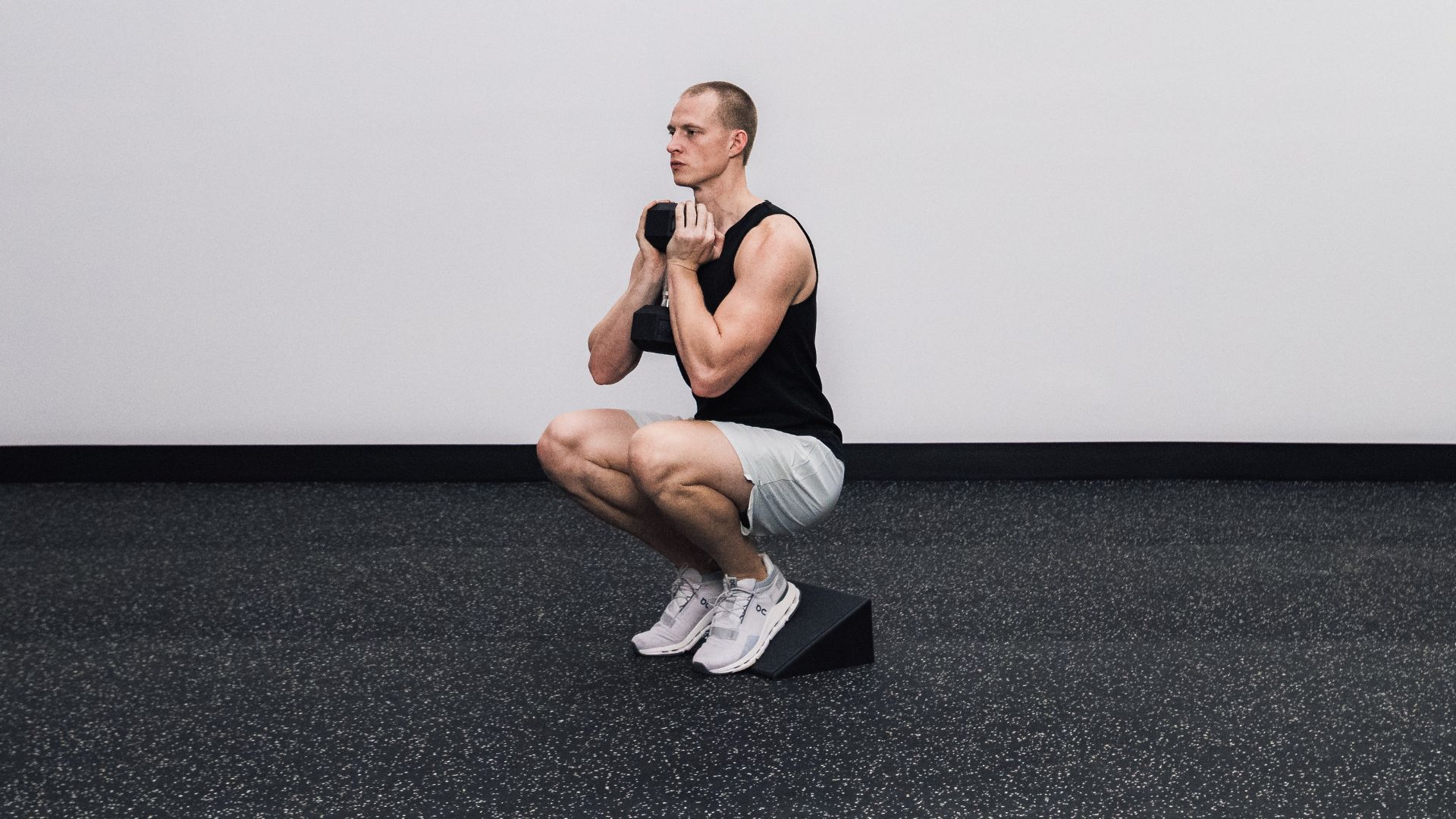 A man in a deep squat holds a dumbbell at chest height with his heels uplifted on a slant board.
