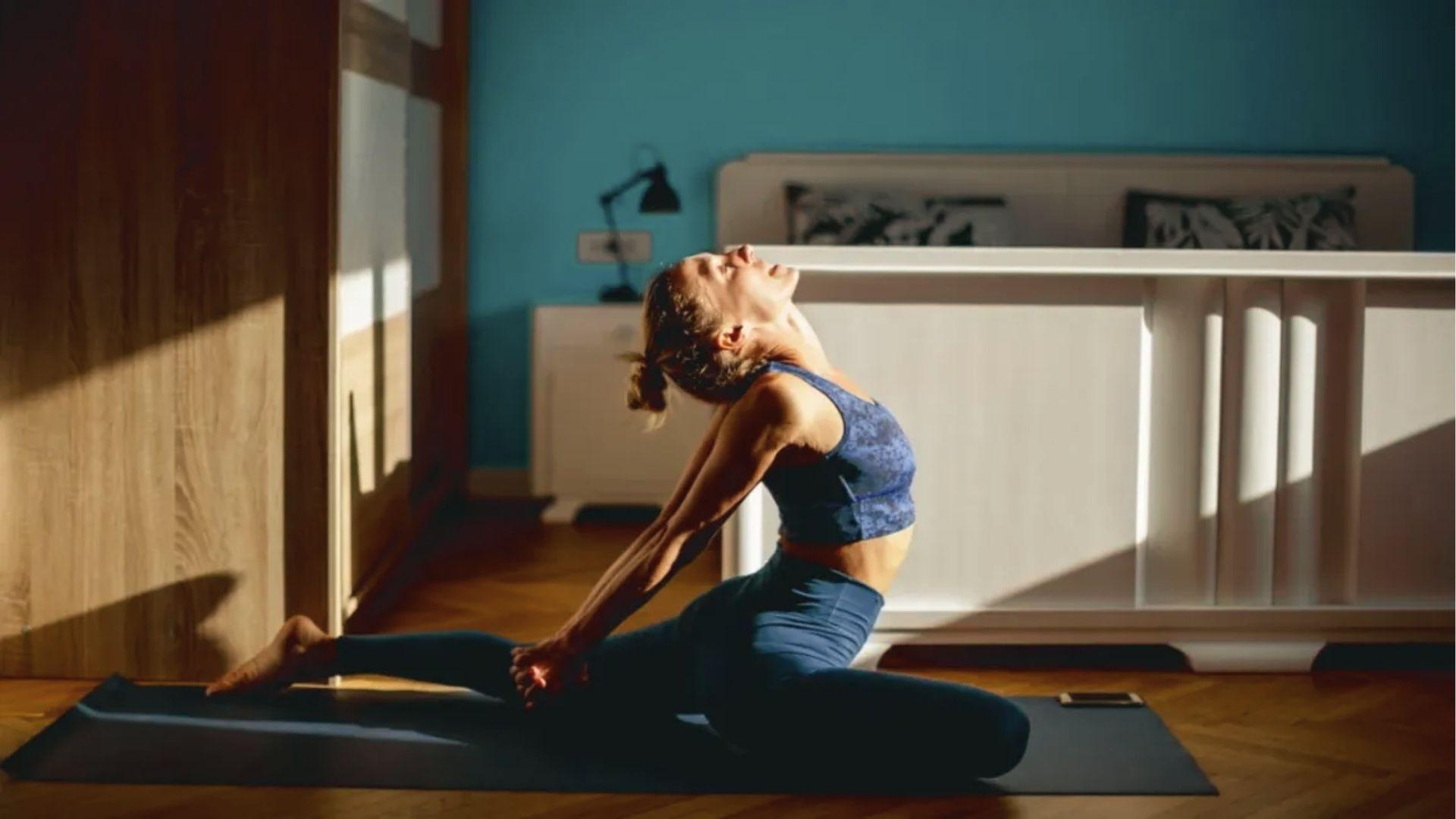A woman in blue activewear does hip flexor lunge stretches on the mat, arching back in the sunlit bedroom.