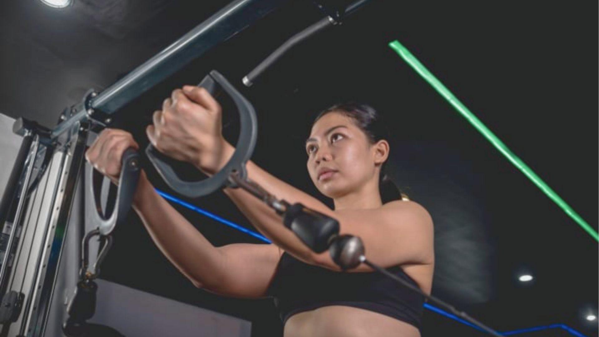 Woman in a black sports bra using cable machine handles, pressing them forward and together in a gym.