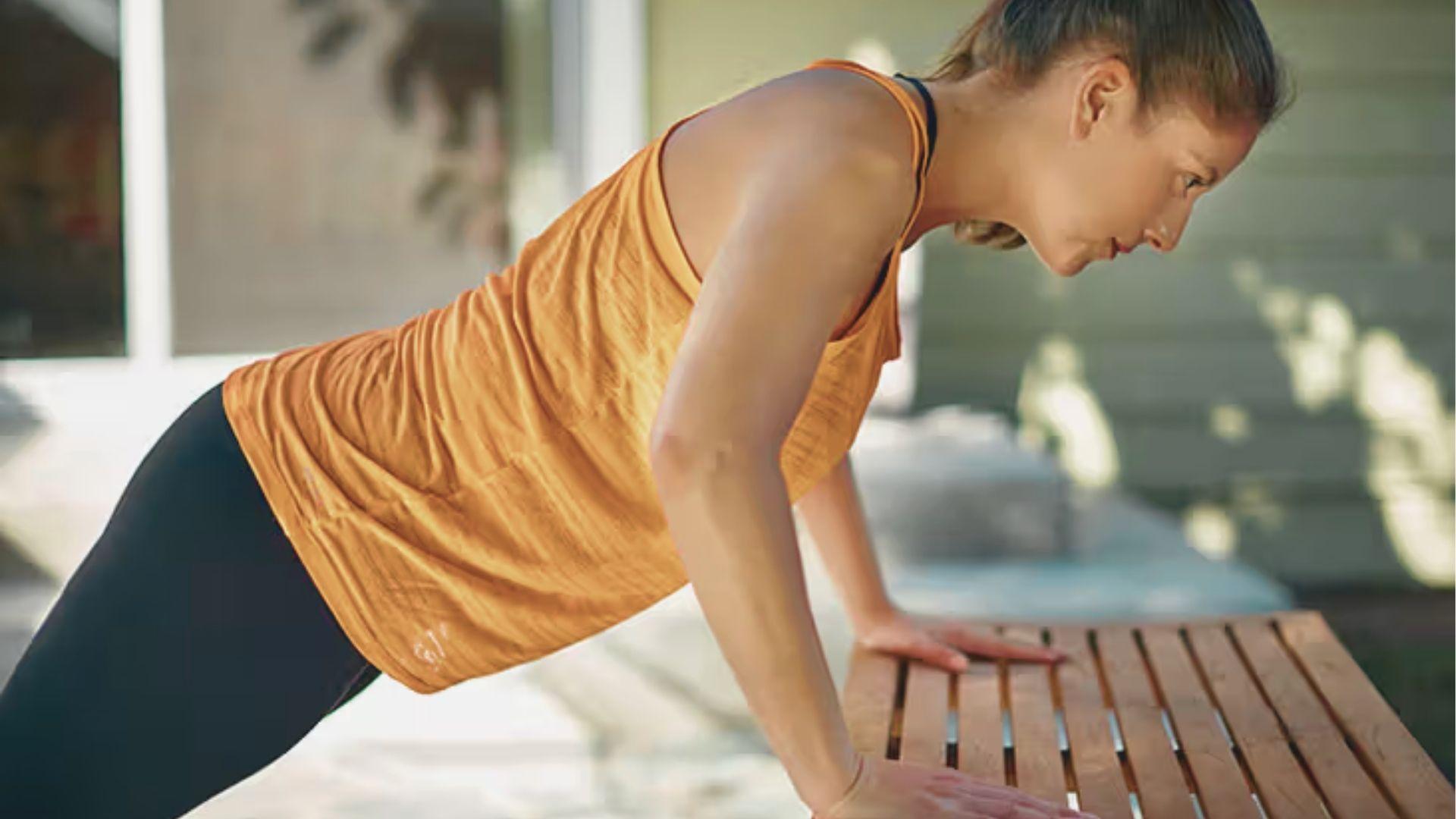 Woman in an orange tank top performing an incline push-up against a wooden table.