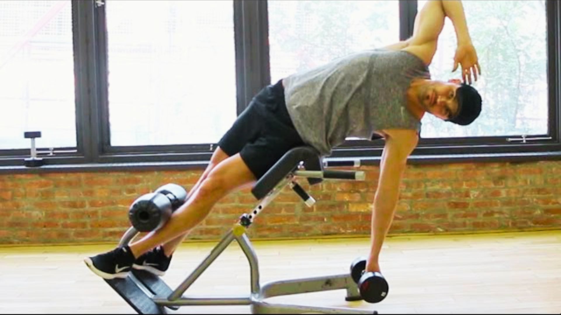 A man in a gray tank top and black shorts does a side bend on a Roman chair while holding a dumbbell.