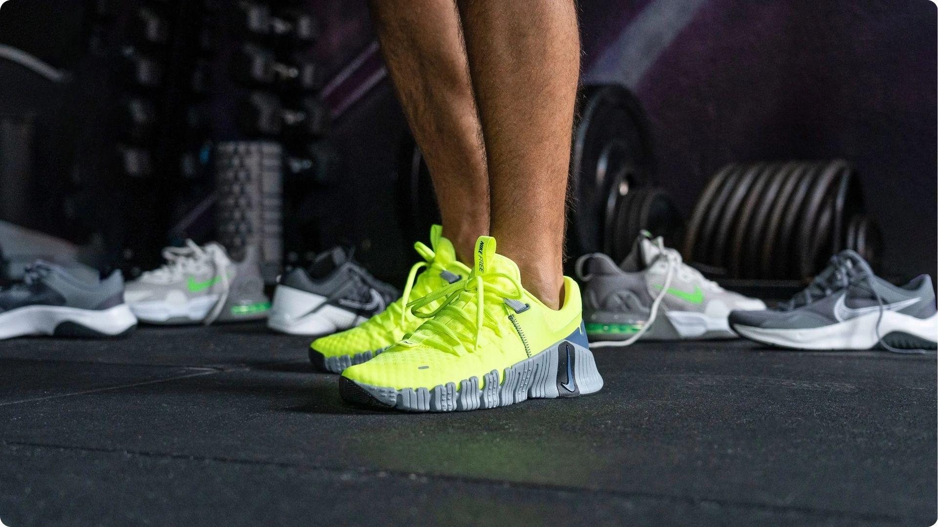 Close-up of a person wearing neon yellow training shoes standing on a gym floor, surrounded by other athletic shoes.