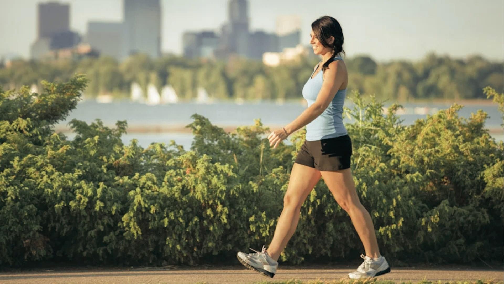 Woman in light blue tank top and black shorts walks briskly along lakeside path with city skyline behind.