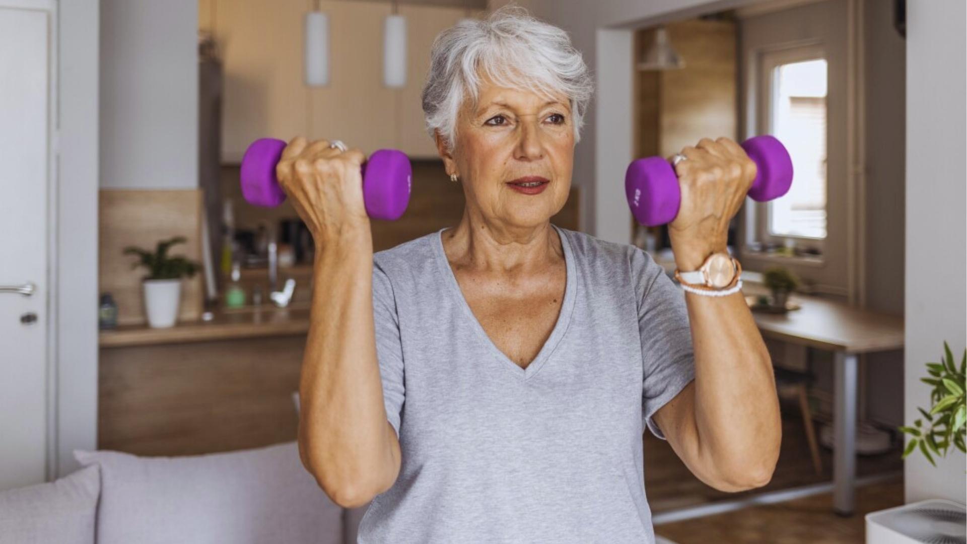 Senior woman with gray hair performs bicep curls with purple dumbbells in a modern kitchen for gentle strength training.