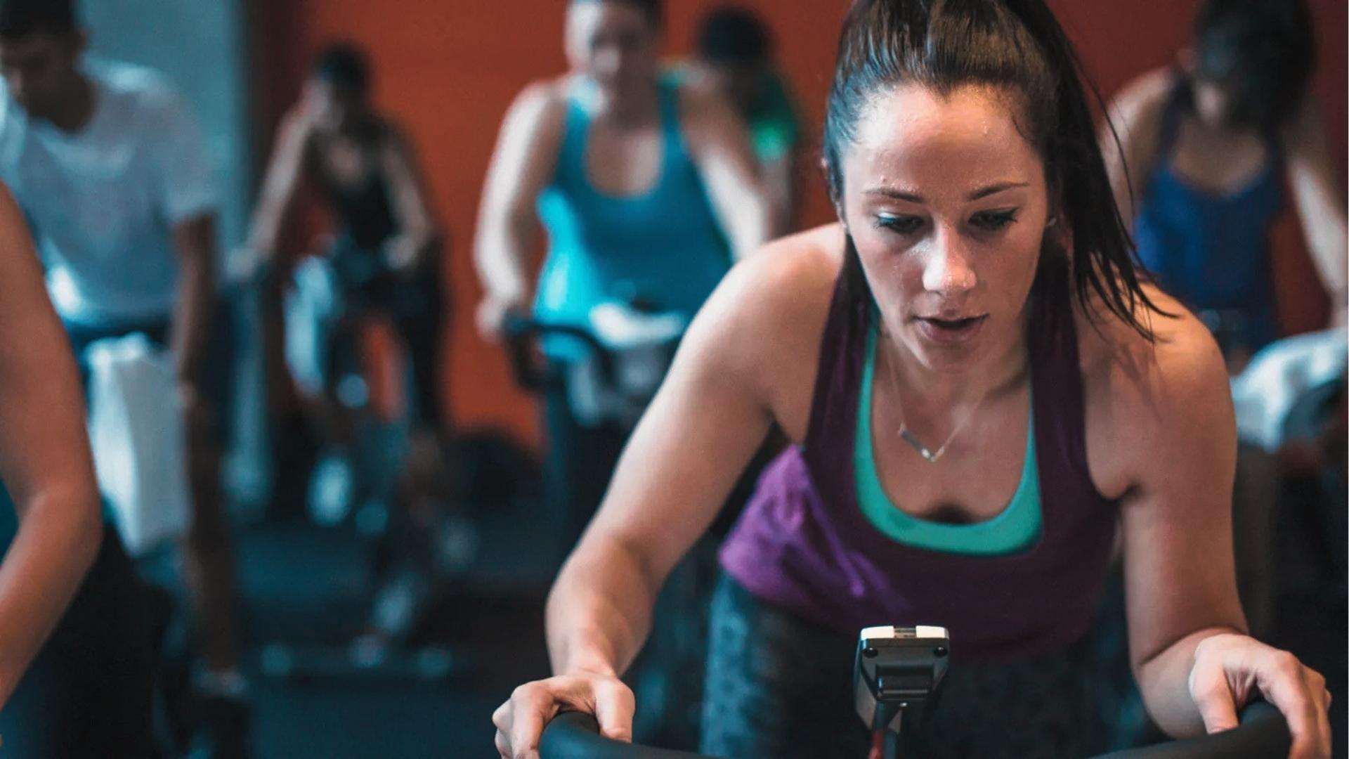 Woman with high ponytail in purple tank top sweats during intense indoor cycling class with other participants blurred behind.