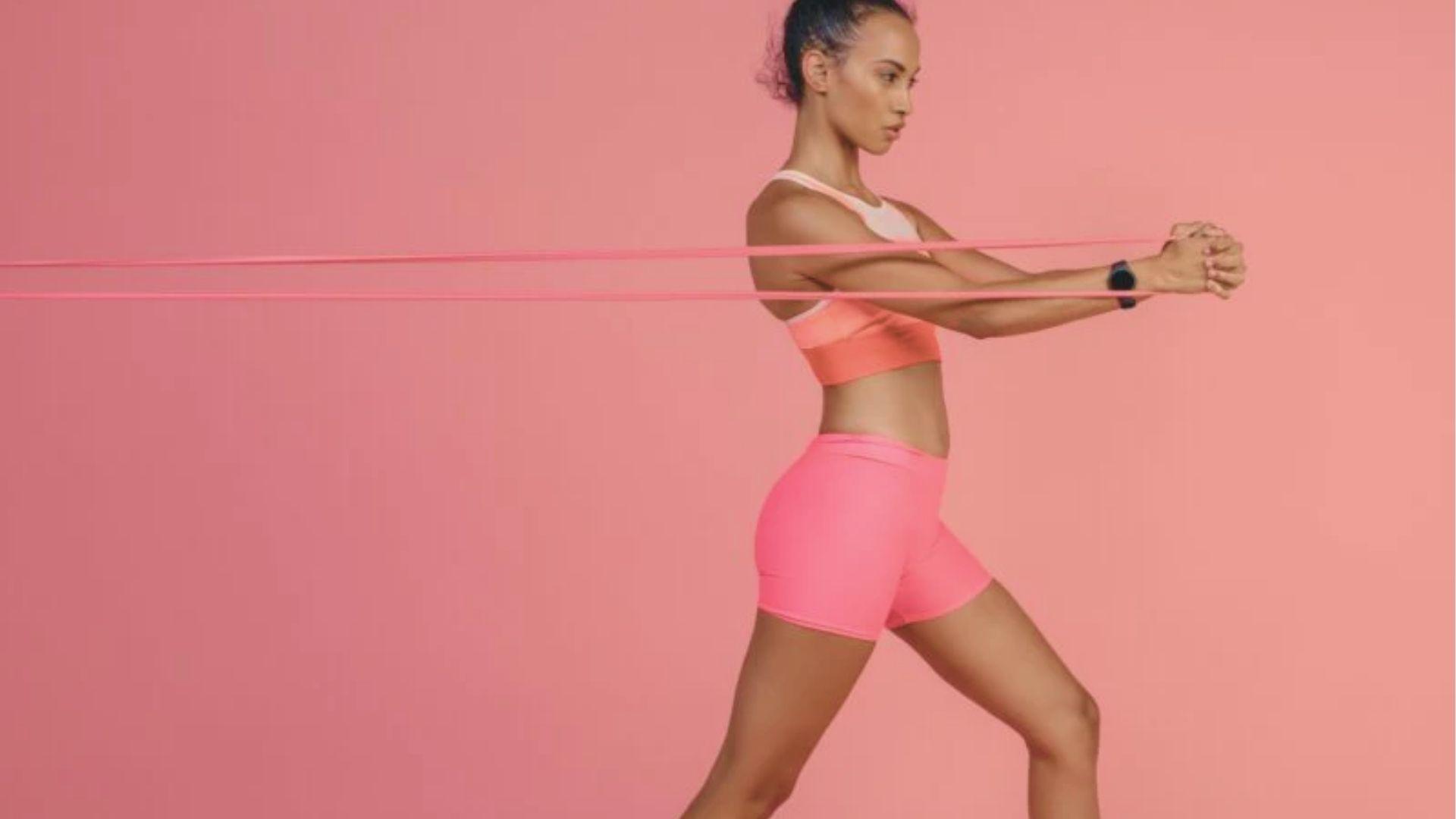 Woman in coral workout set standing against a pink background, pressing a resistance band forward with both hands.