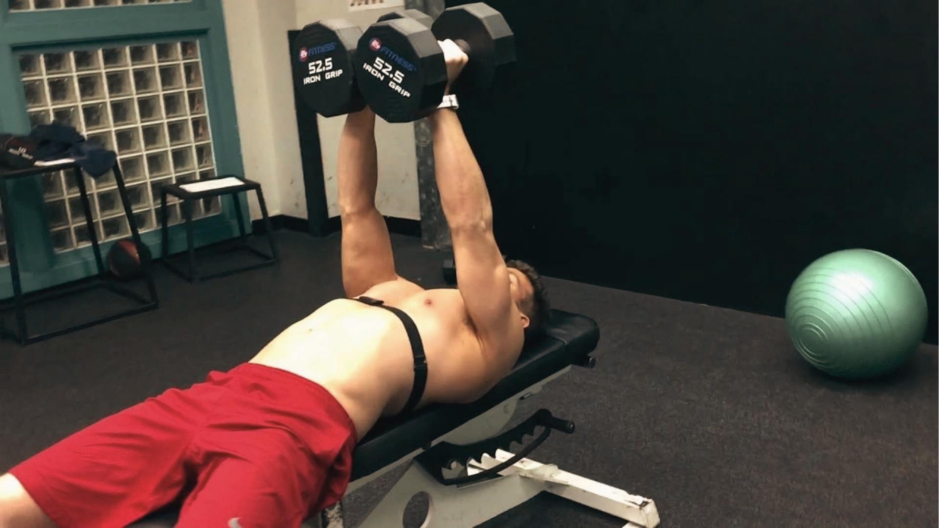 Shirtless man in red shorts lies on a flat bench pressing 52.5 lb dumbbells together for close grip chest press.