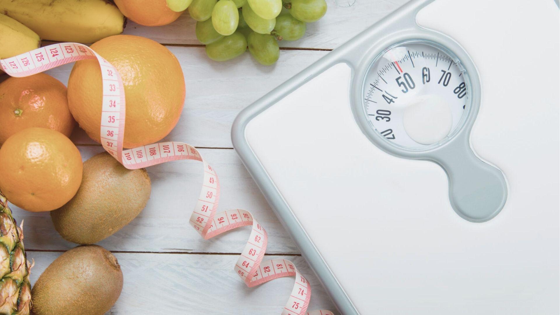 Flat lay of fresh fruit, measuring tape, and a bathroom scale on a white wood surface.