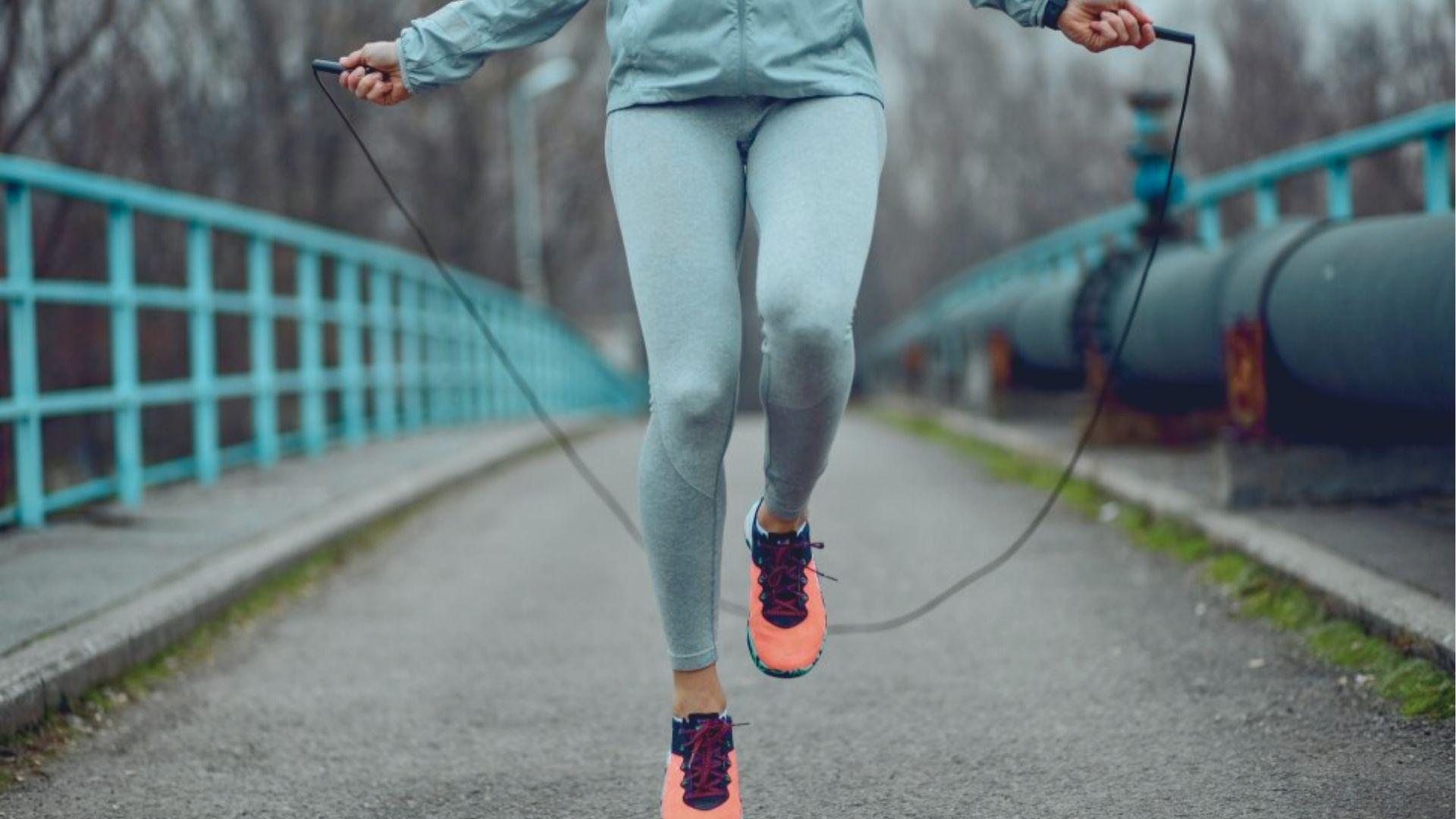 Person in gray activewear jump ropes outdoors on a bridge, mid-air with rope under feet.