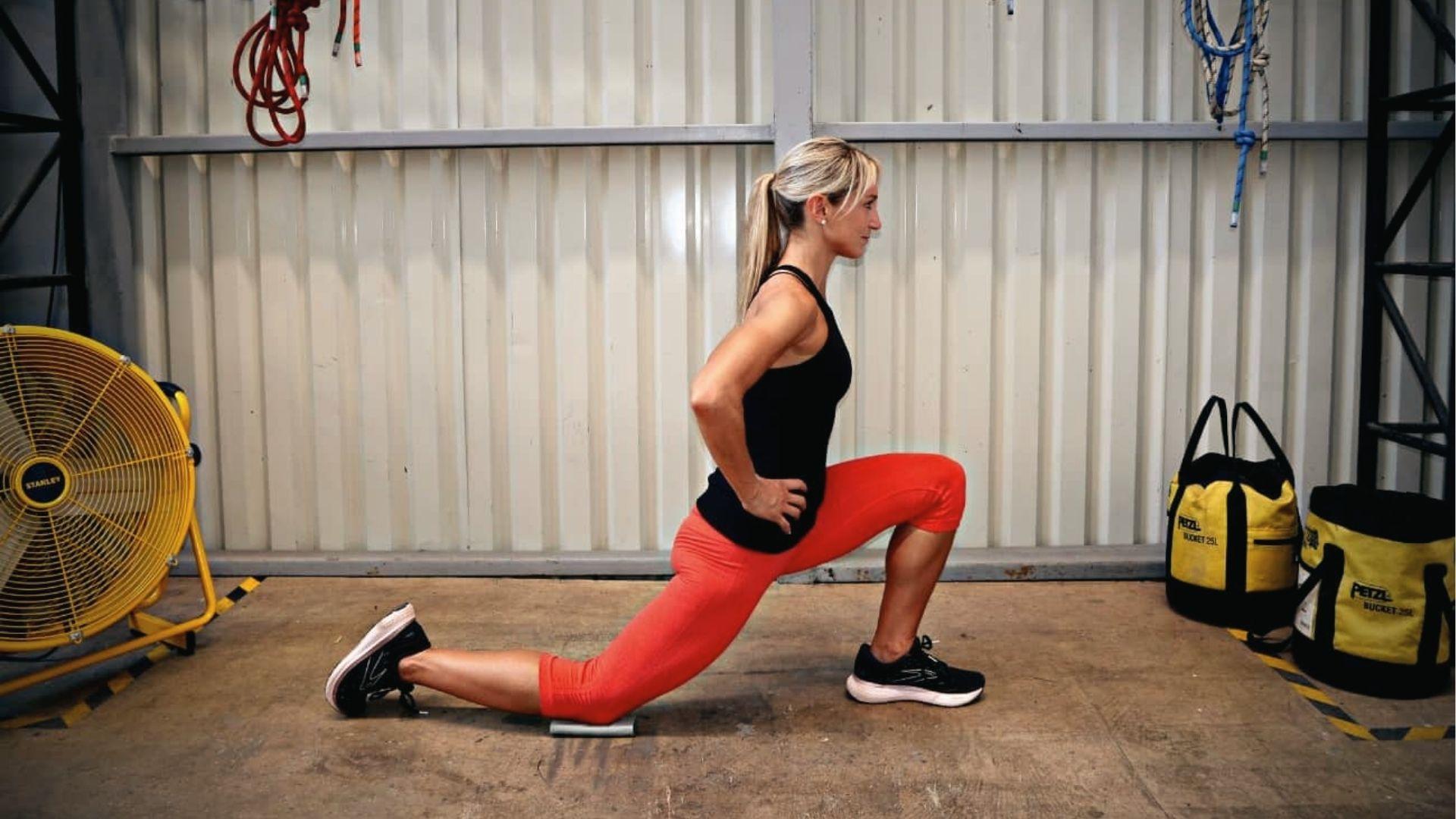 A woman in red leggings performs hip flexor lunge stretches with a knee pad in the gym, hands on hips.