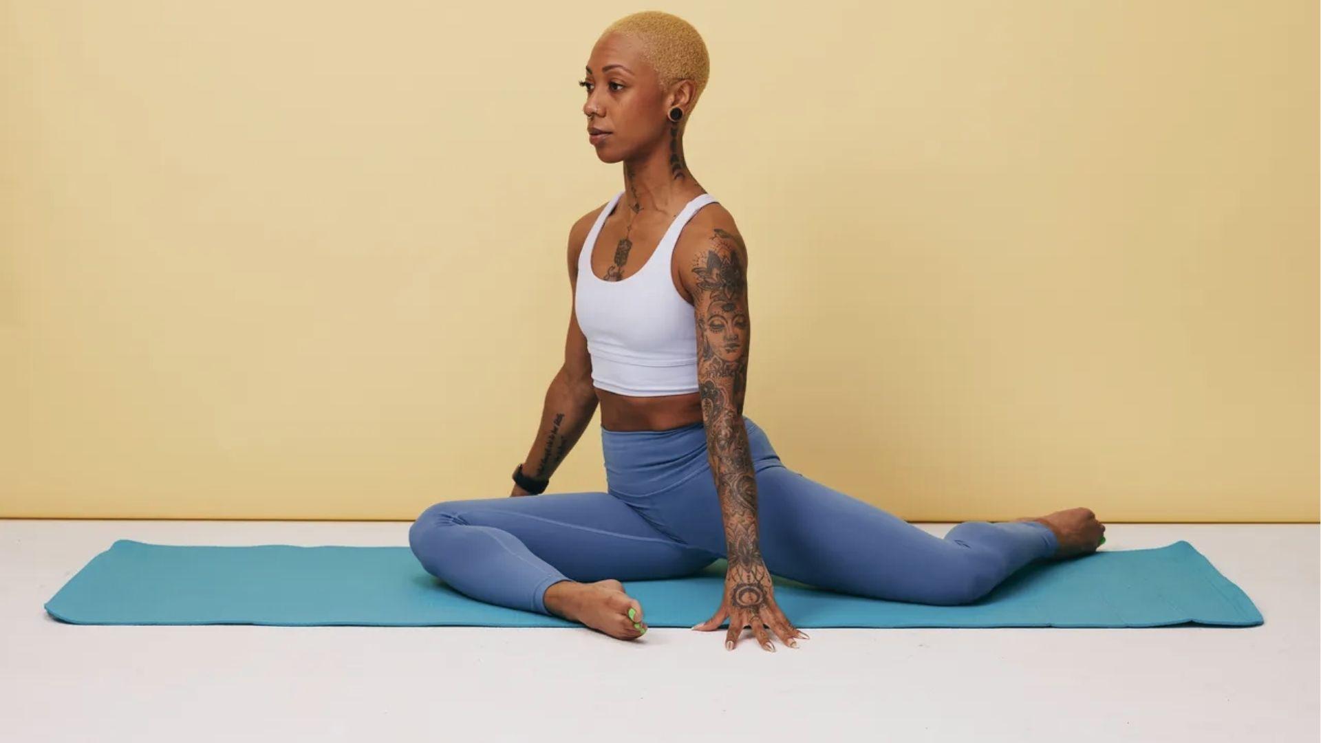 A tattooed woman in white top holds a pigeon pose variation to stretch hip flexors on a blue yoga mat.