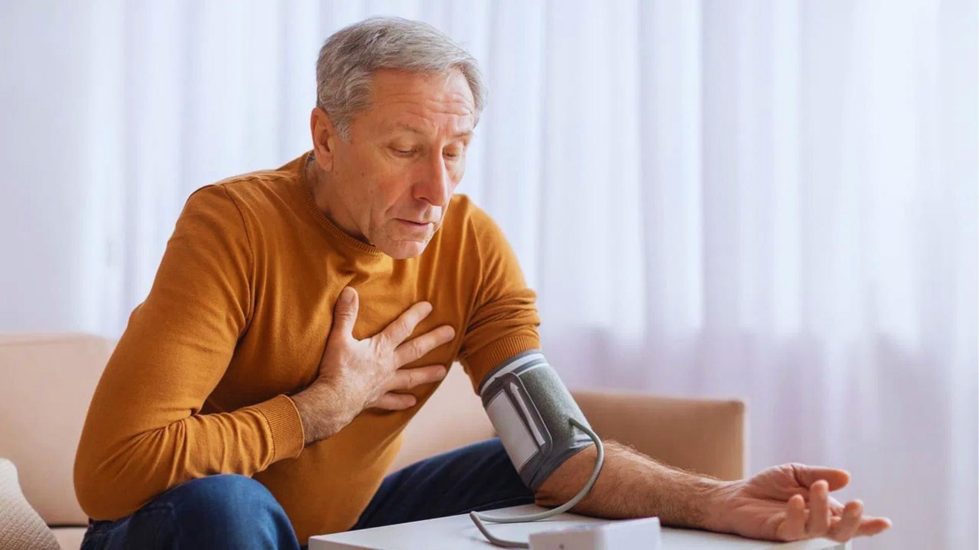 Concerned senior man in mustard sweater checks blood pressure at home while holding his chest, monitoring heart health.