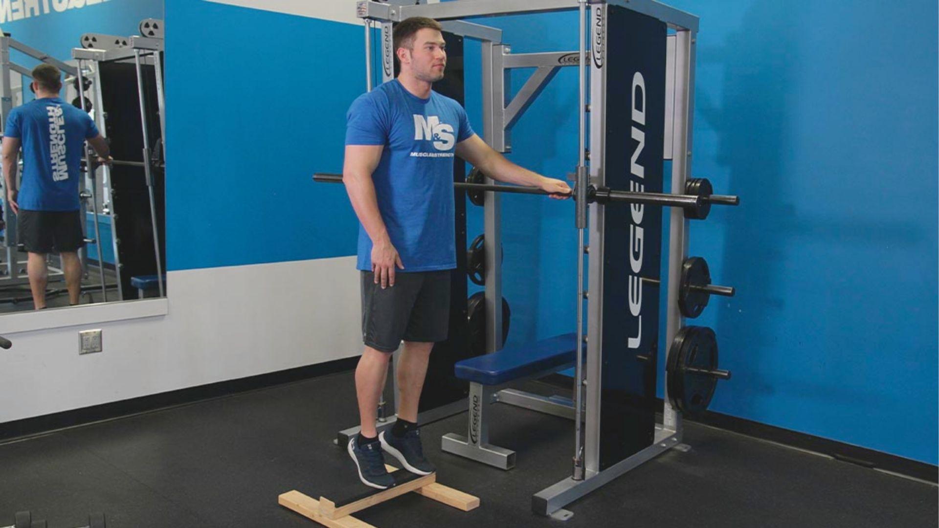 Man in blue Muscle & Strength shirt stands on a wooden calf block without weights, hand on Smith machine for support.