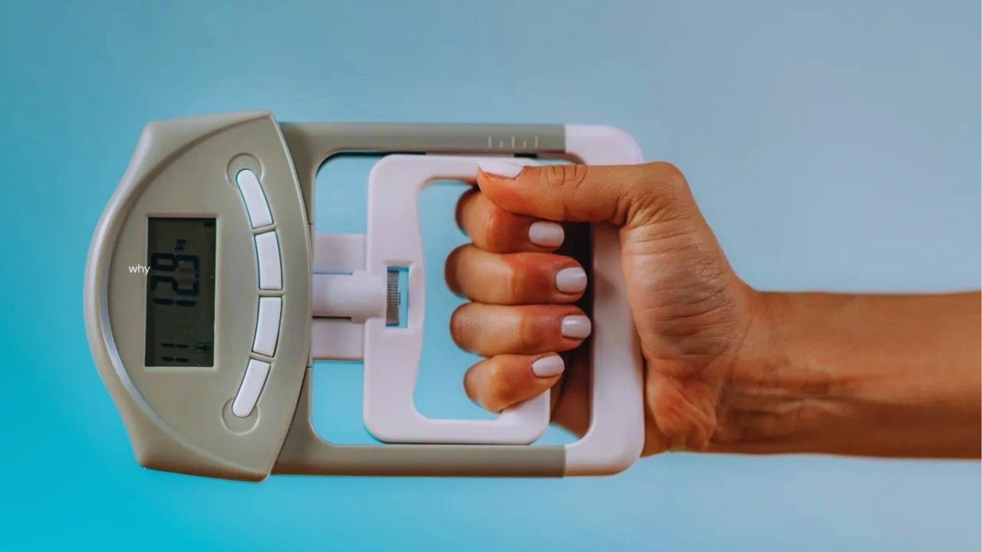 Woman’s hand with light pink manicure grips a digital hand dynamometer reading on a bright blue background.