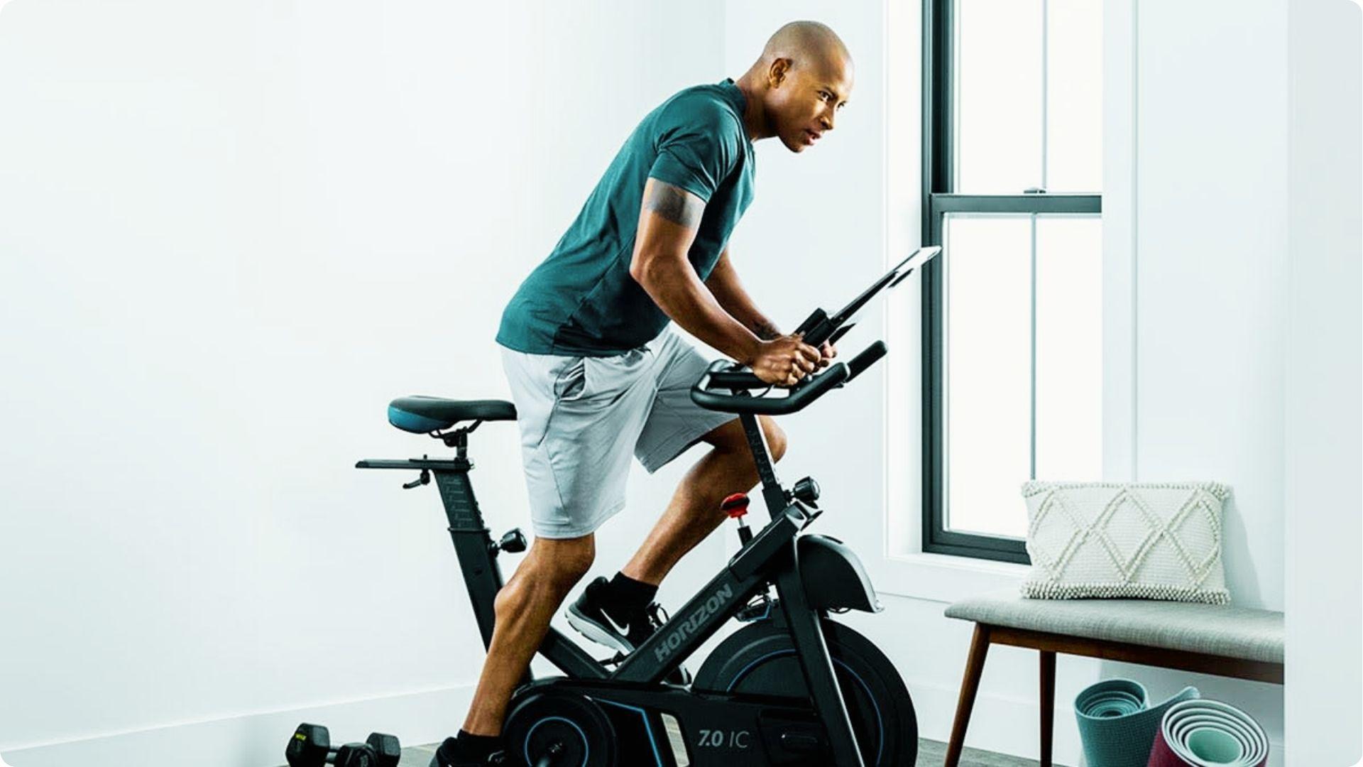 A man rides an indoor cycling bike by a window in a bright, minimalist room.