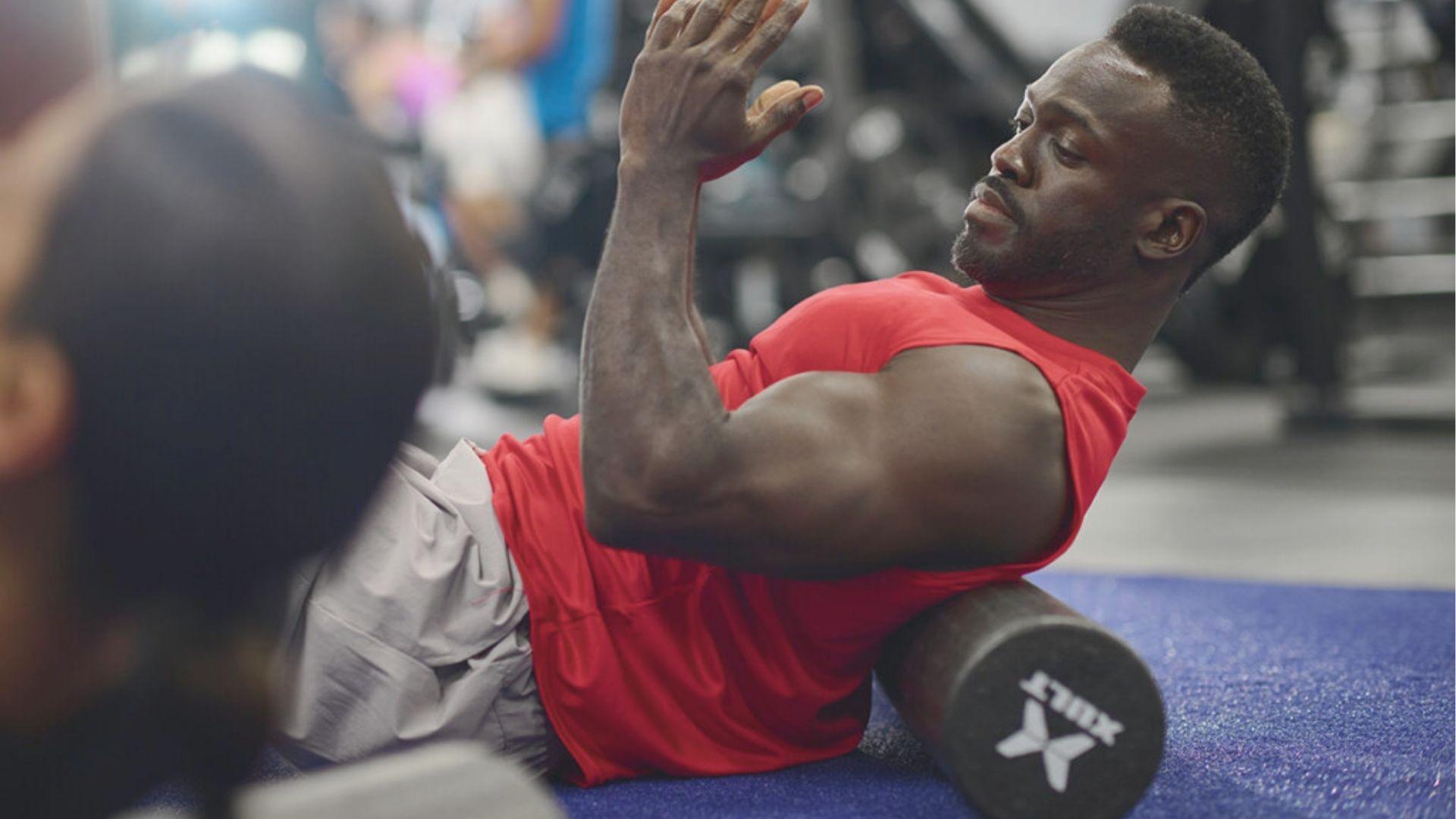 Man in red tank top lies on a foam roller at the gym, targeting his upper back muscles.