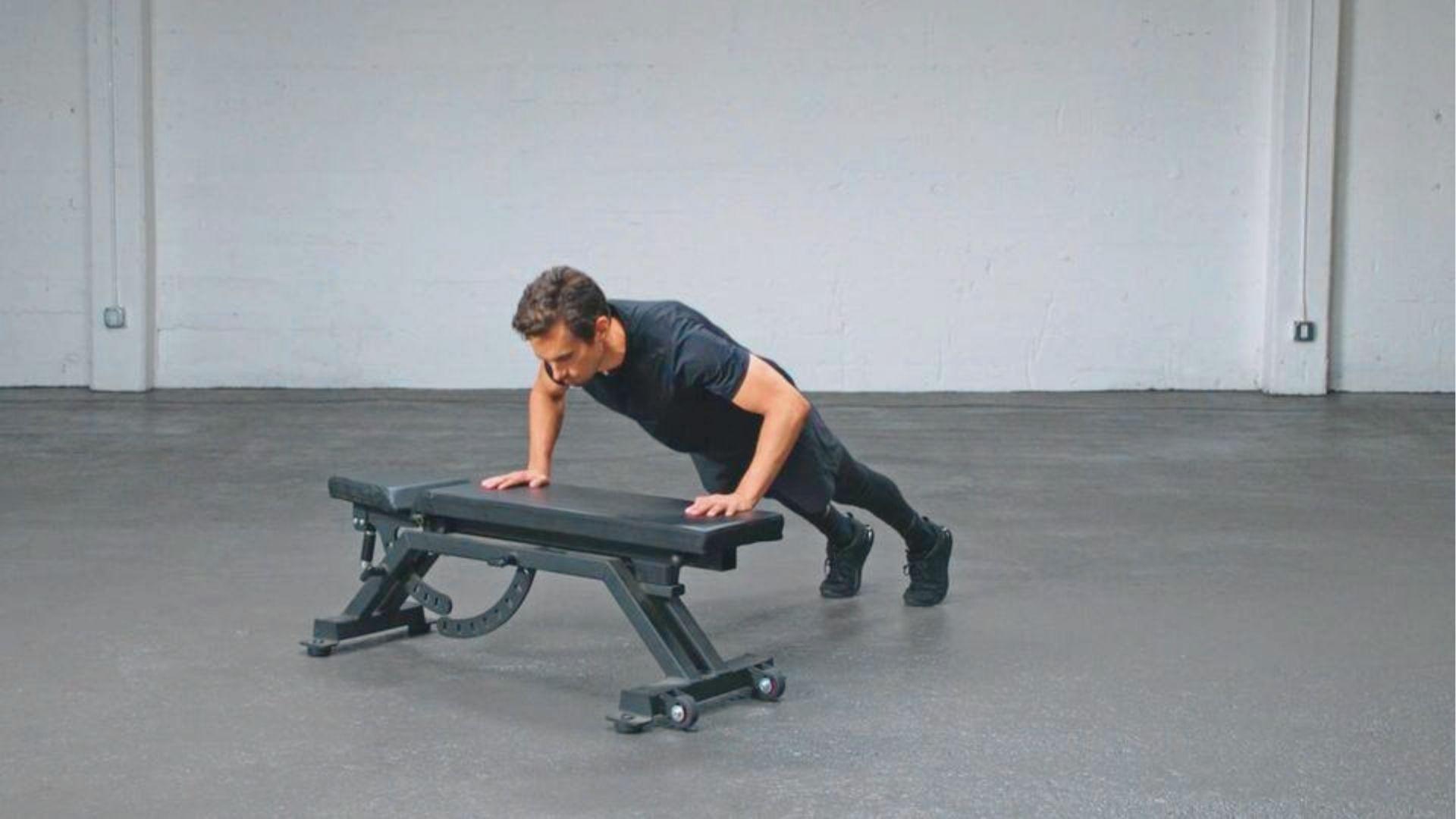 Man in black athletic wear performing an incline negative push up on a gym bench.