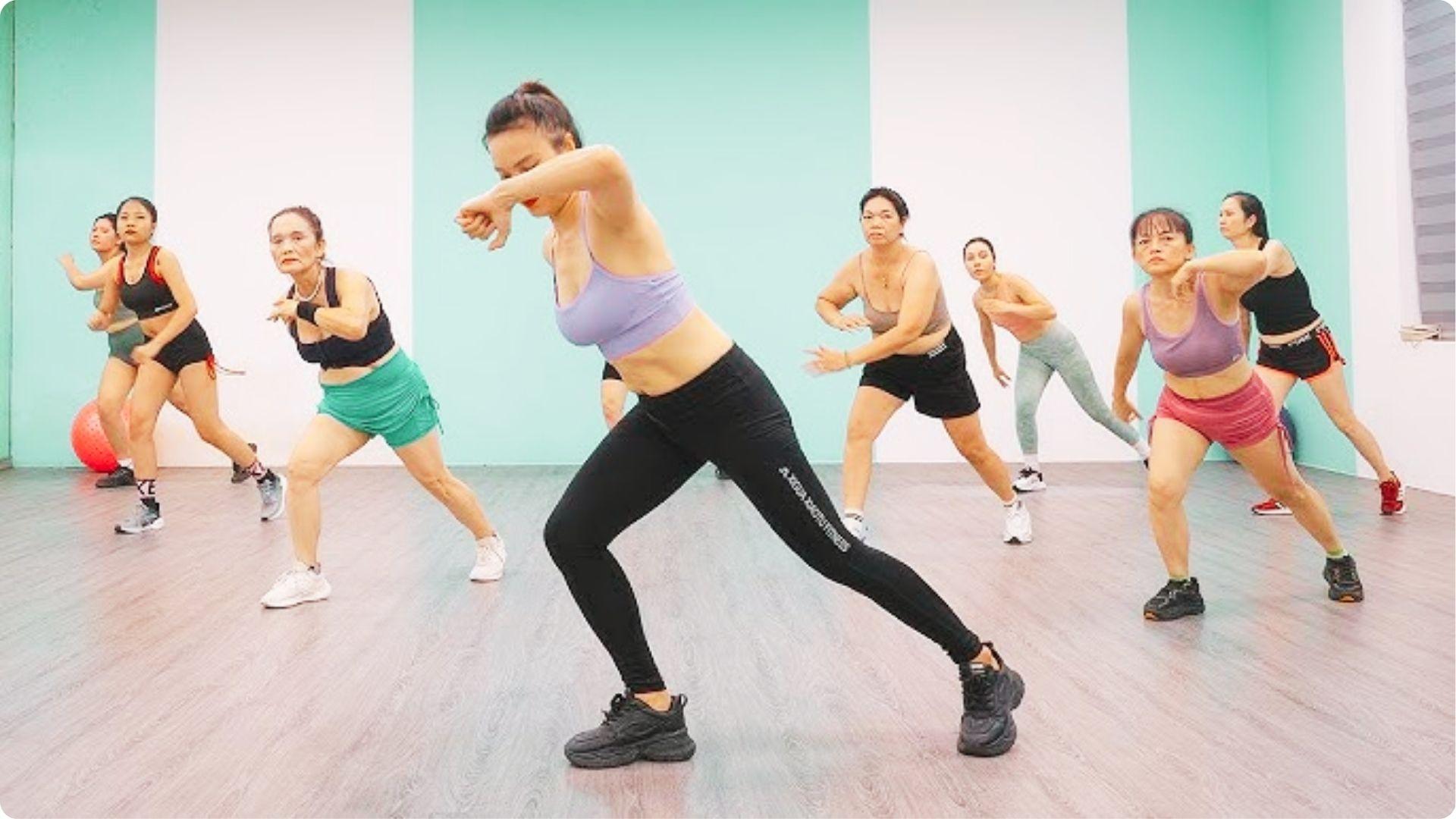 A group of women follows an instructor in a dance fitness class with side-step movements in a bright studio.