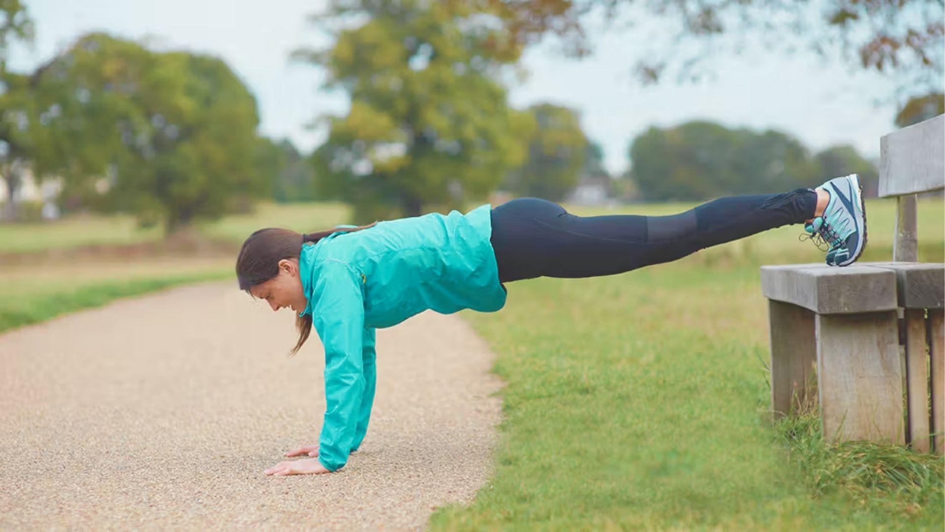 Woman in a teal jacket holding a decline push-up position with feet elevated on a park bench.