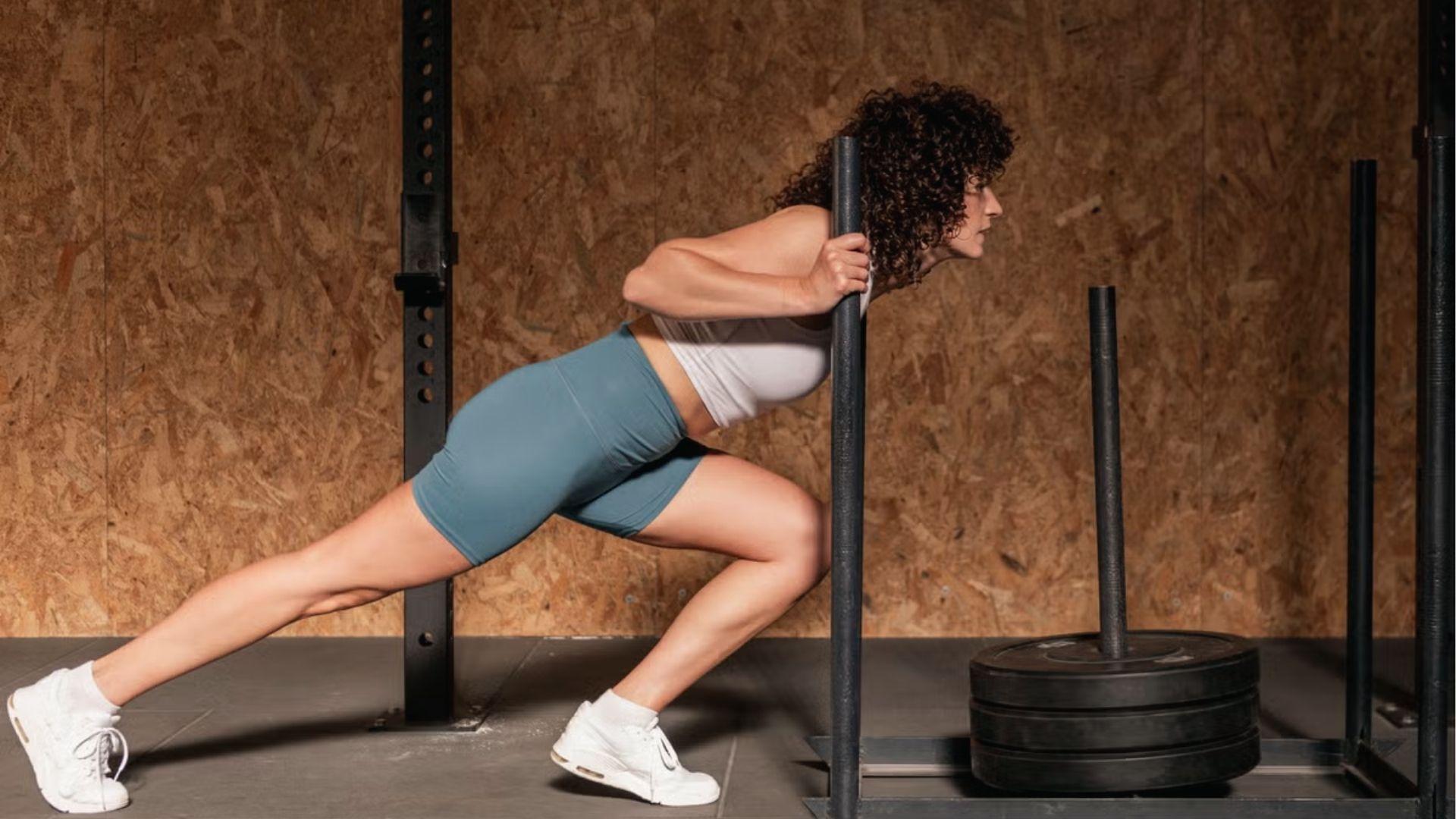A woman in teal shorts and white top performs low-angle sled push, gripping handles with strong forward lean in the gym.