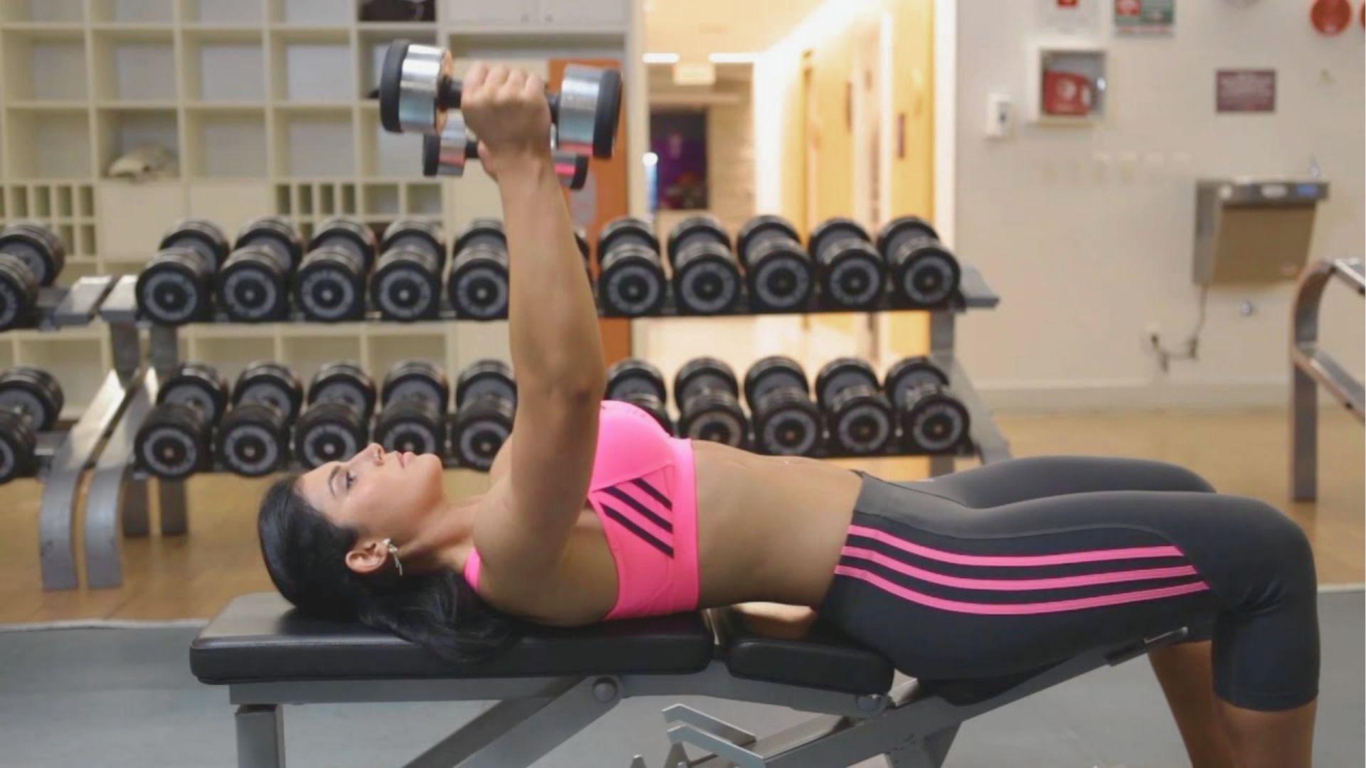 Woman in a pink sports bra lying on a flat bench, pressing two dumbbells straight up over her chest.