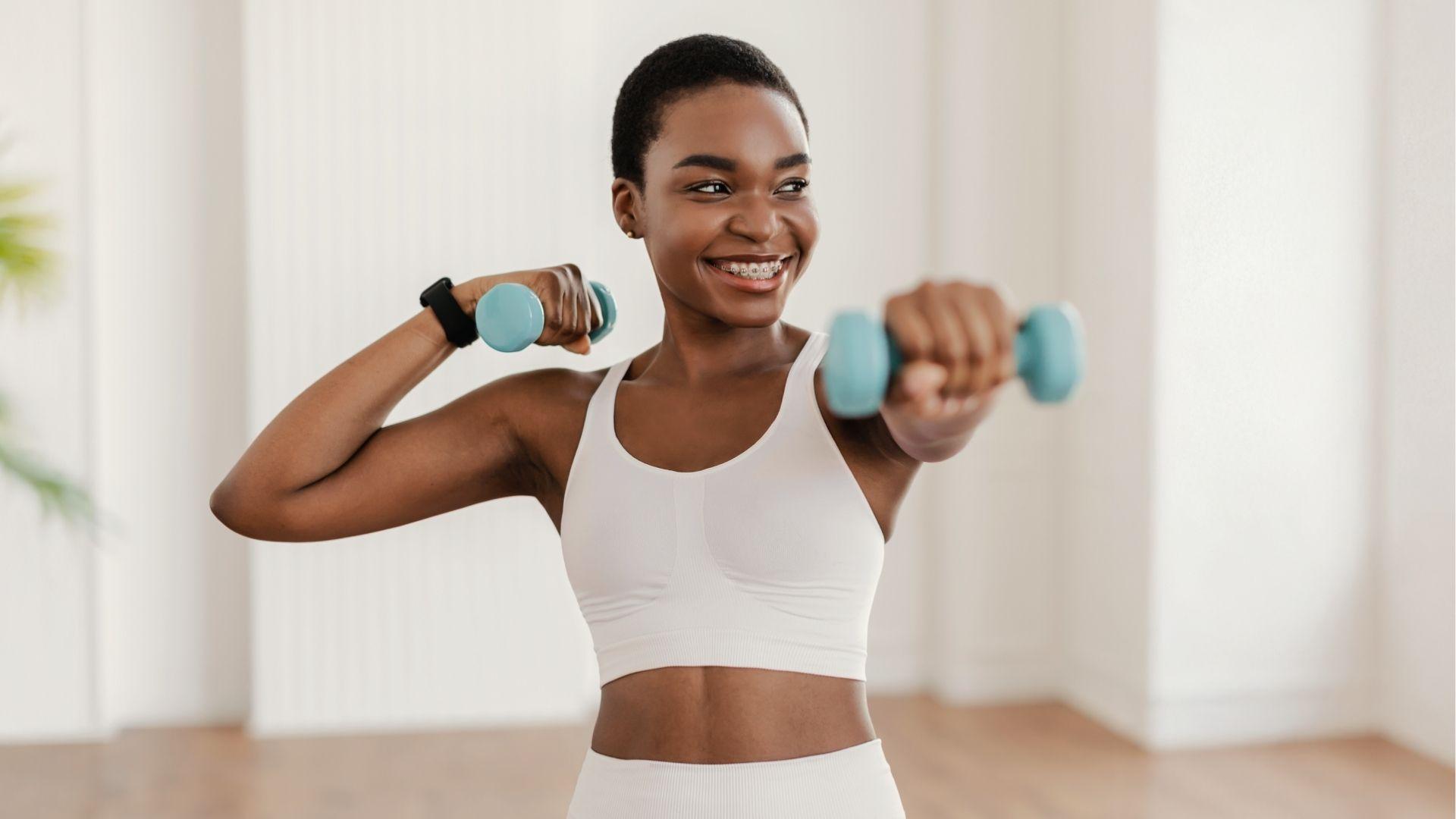 Smiling woman lifts light dumbbells indoors adding strength training to bodyweight calisthenics days