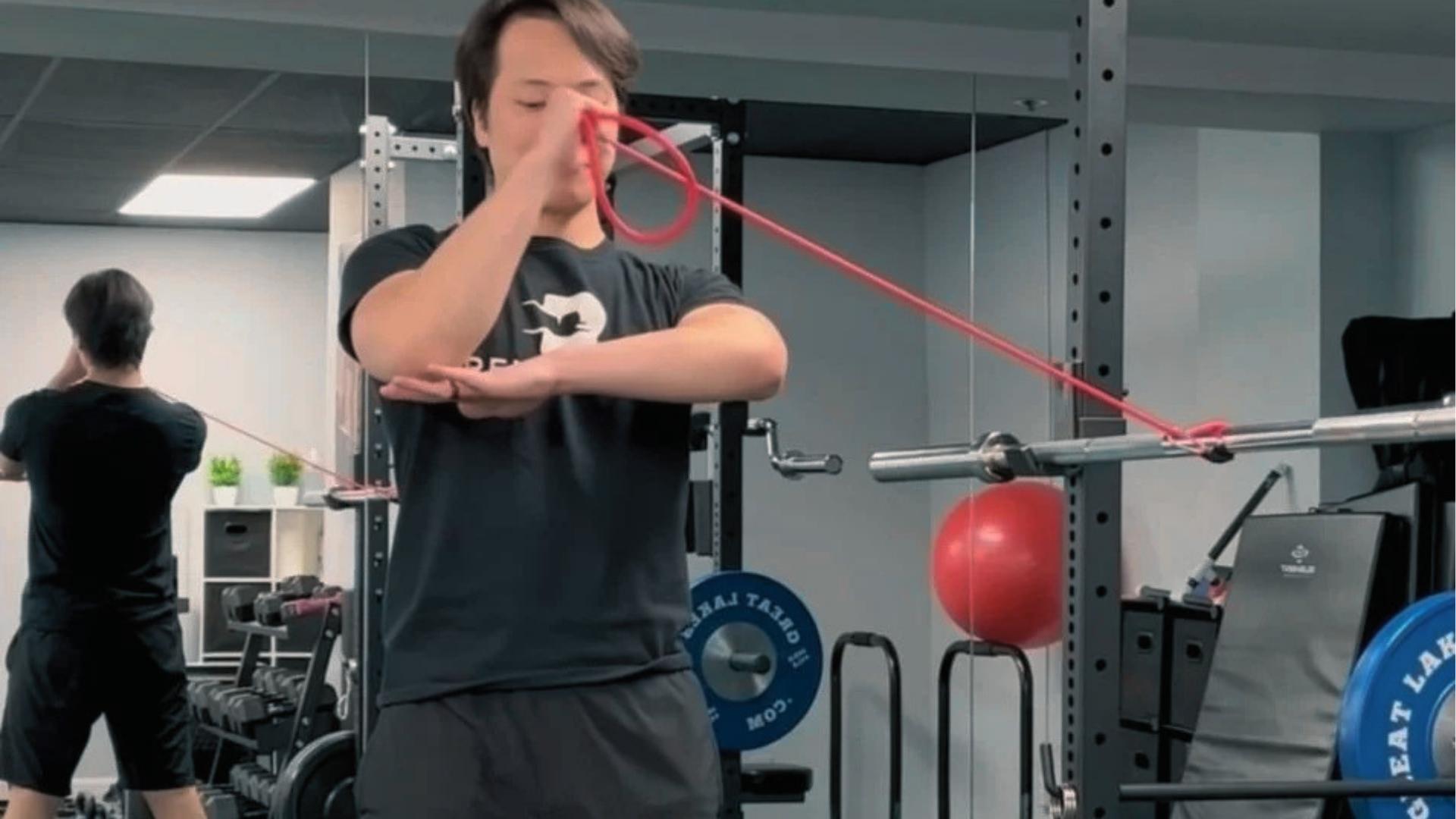 Man in gym performs rotation with red resistance band anchored to rack, elbow bent and stabilized.