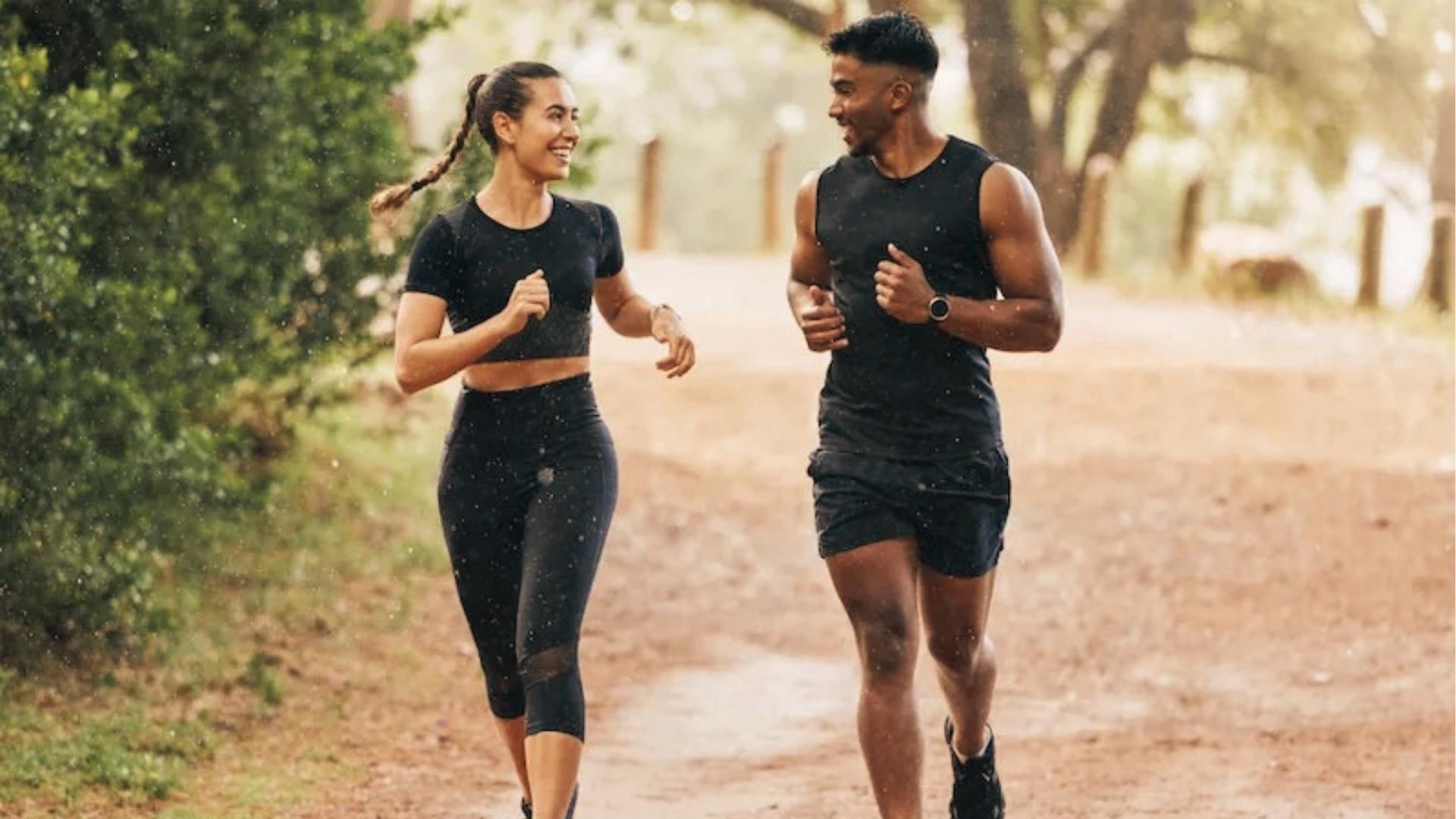 Smiling man and woman in black athletic wear jog together on dirt trail surrounded by trees, tracking steps.