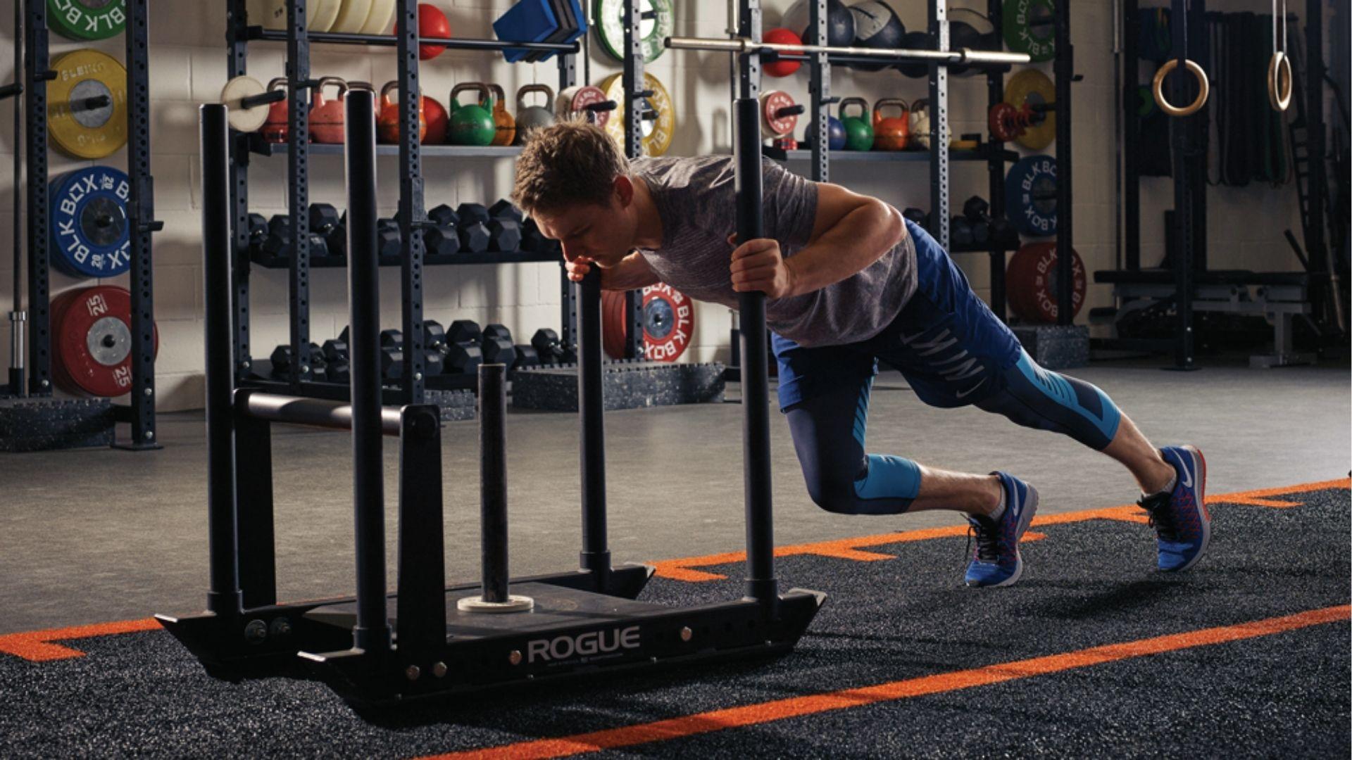 A man in a gray shirt drives a Rogue sled across a turf track in the gym, demonstrating explosive strength and sled push form.
