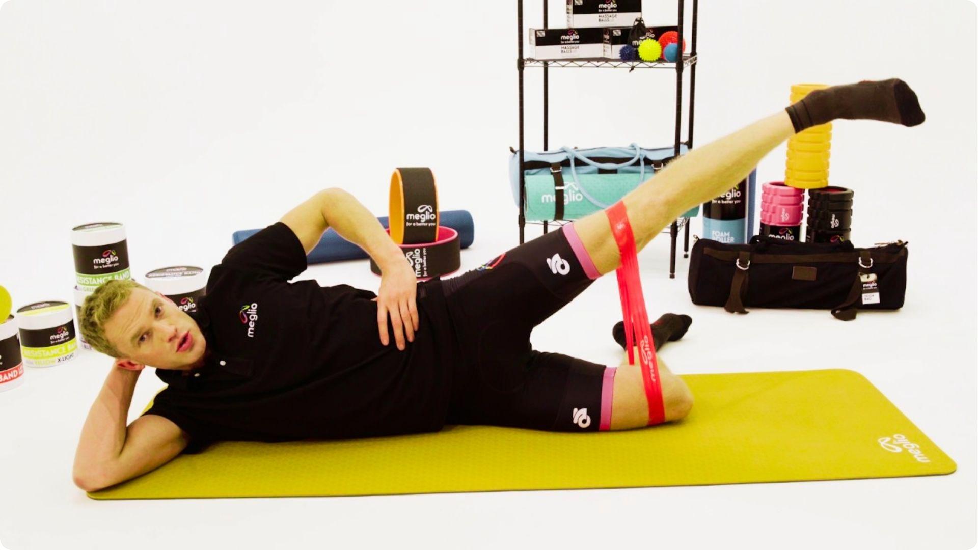 Man in black athletic wear lies on a yellow mat doing a side-lying leg raise with a red resistance band around his thighs.