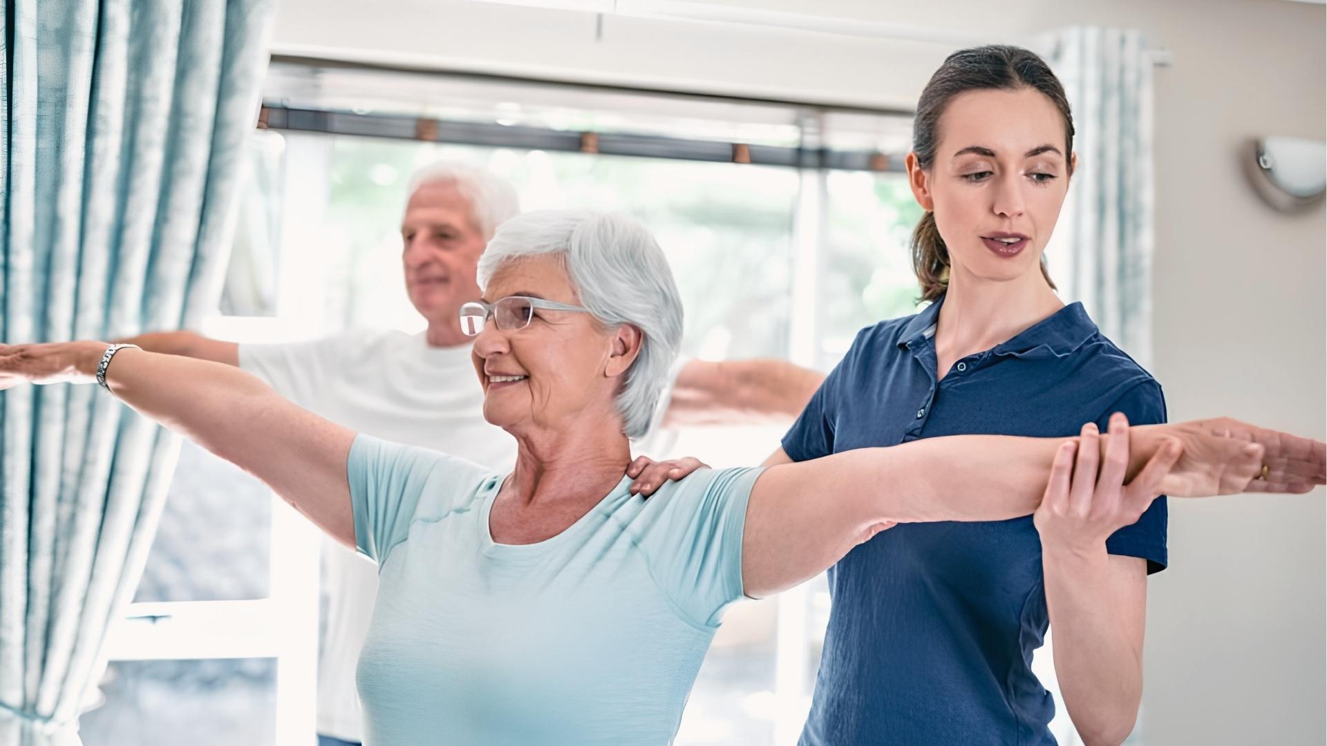 Physical therapist guides senior women through gentle arm stretches during rehab sessions, with elderly men exercising behind them.