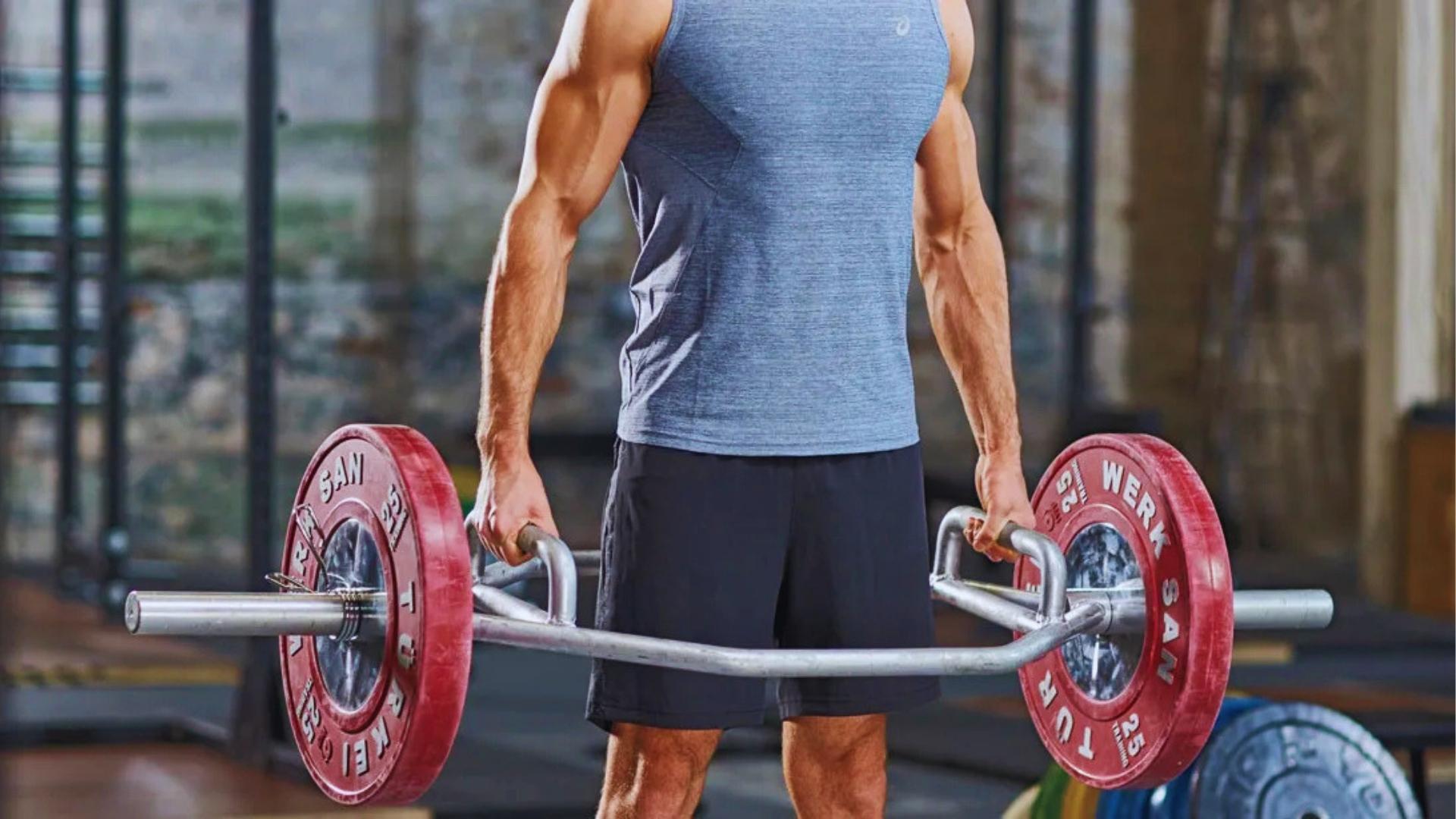 Man in blue sleeveless shirt and black shorts stands tall holding a loaded hex bar with red plates in an industrial gym.
