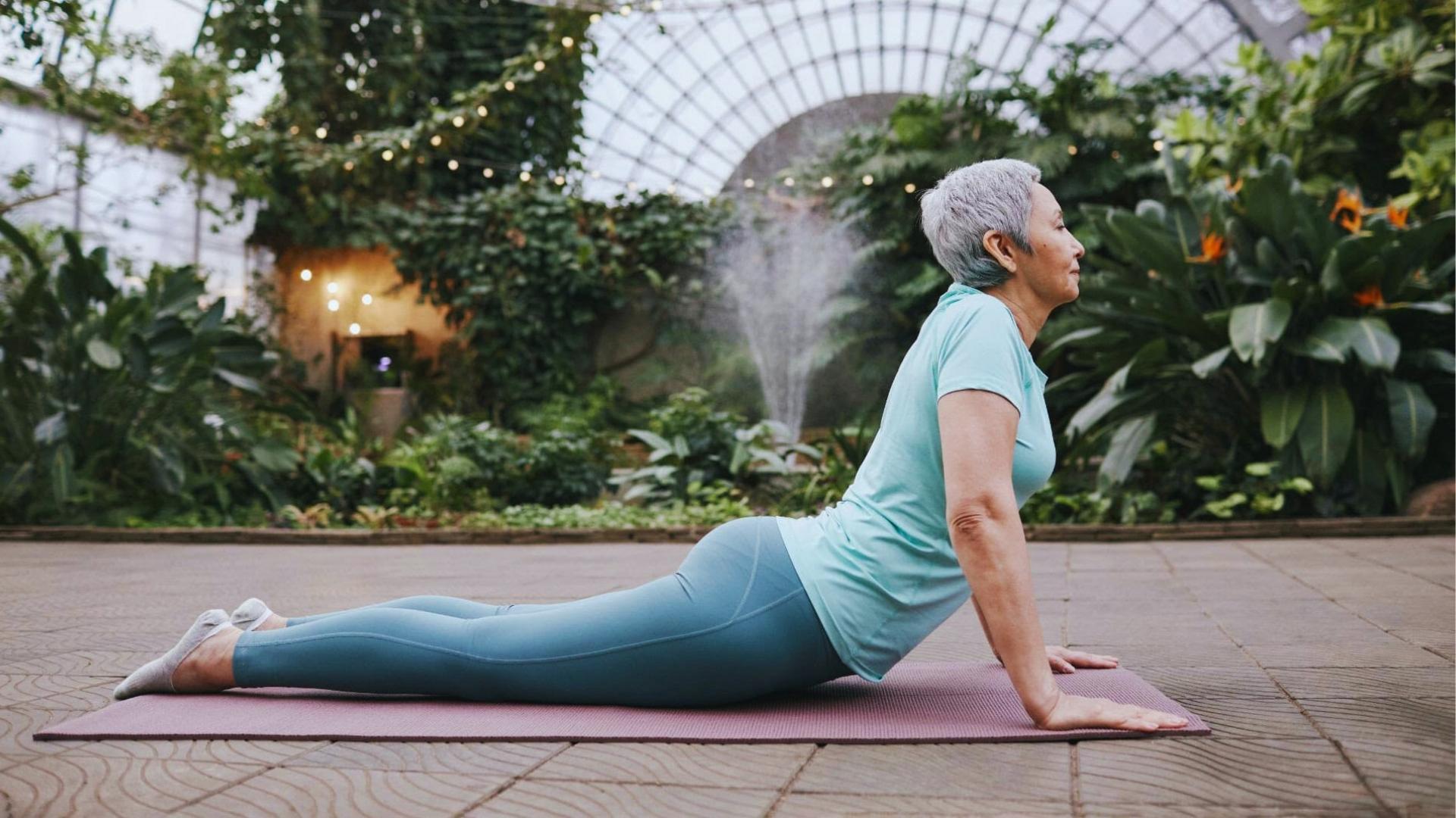 Older woman in teal T-shirt does cobra yoga pose on mat in greenhouse, gently stretching spine and chest.