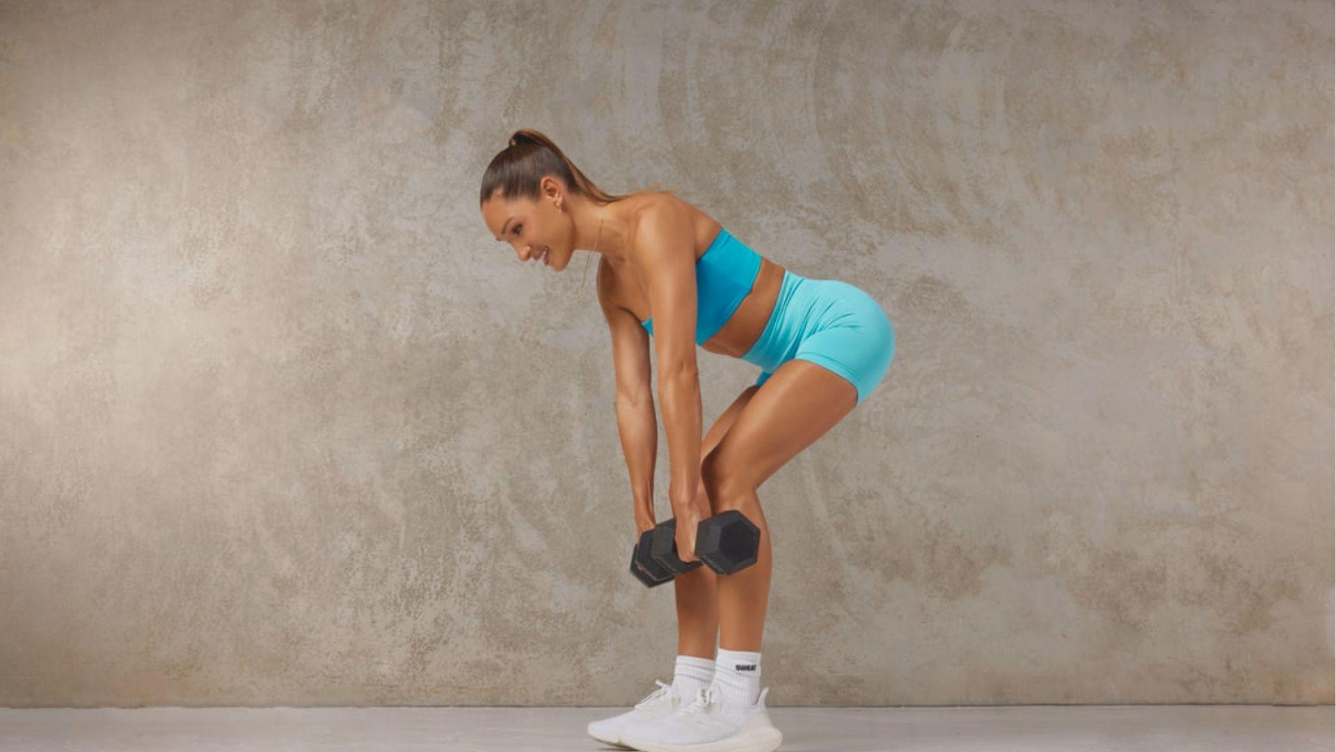 Woman in blue sports bra and shorts performing dumbbell RDL against a beige studio backdrop, mid-hinge with weights at knees.