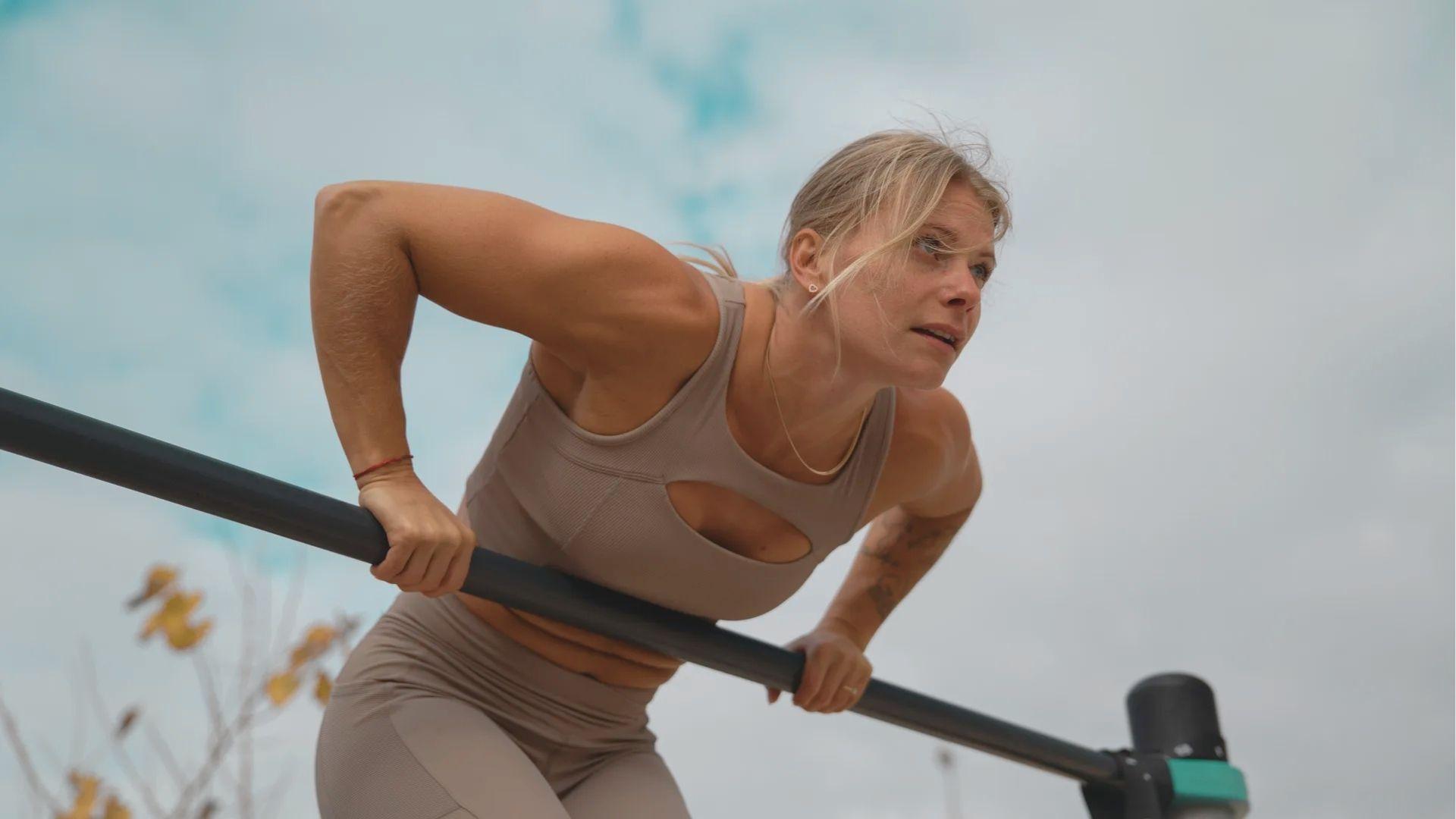 Athletic woman in beige sports bra and leggings performing a muscle-up transition on an outdoor bar.