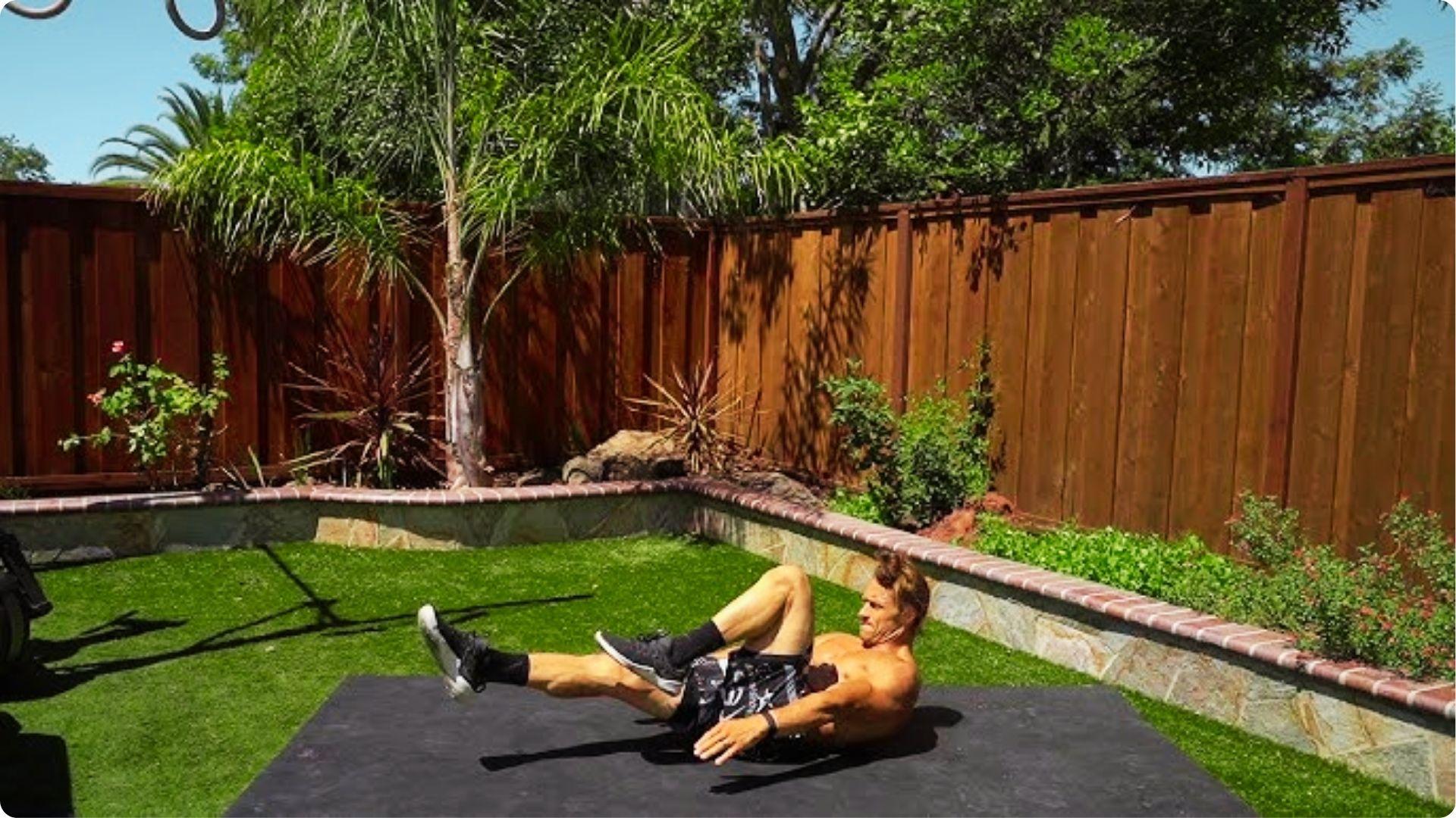A man outdoors does a hollow body rock variation on a black mat in a sunny backyard.
