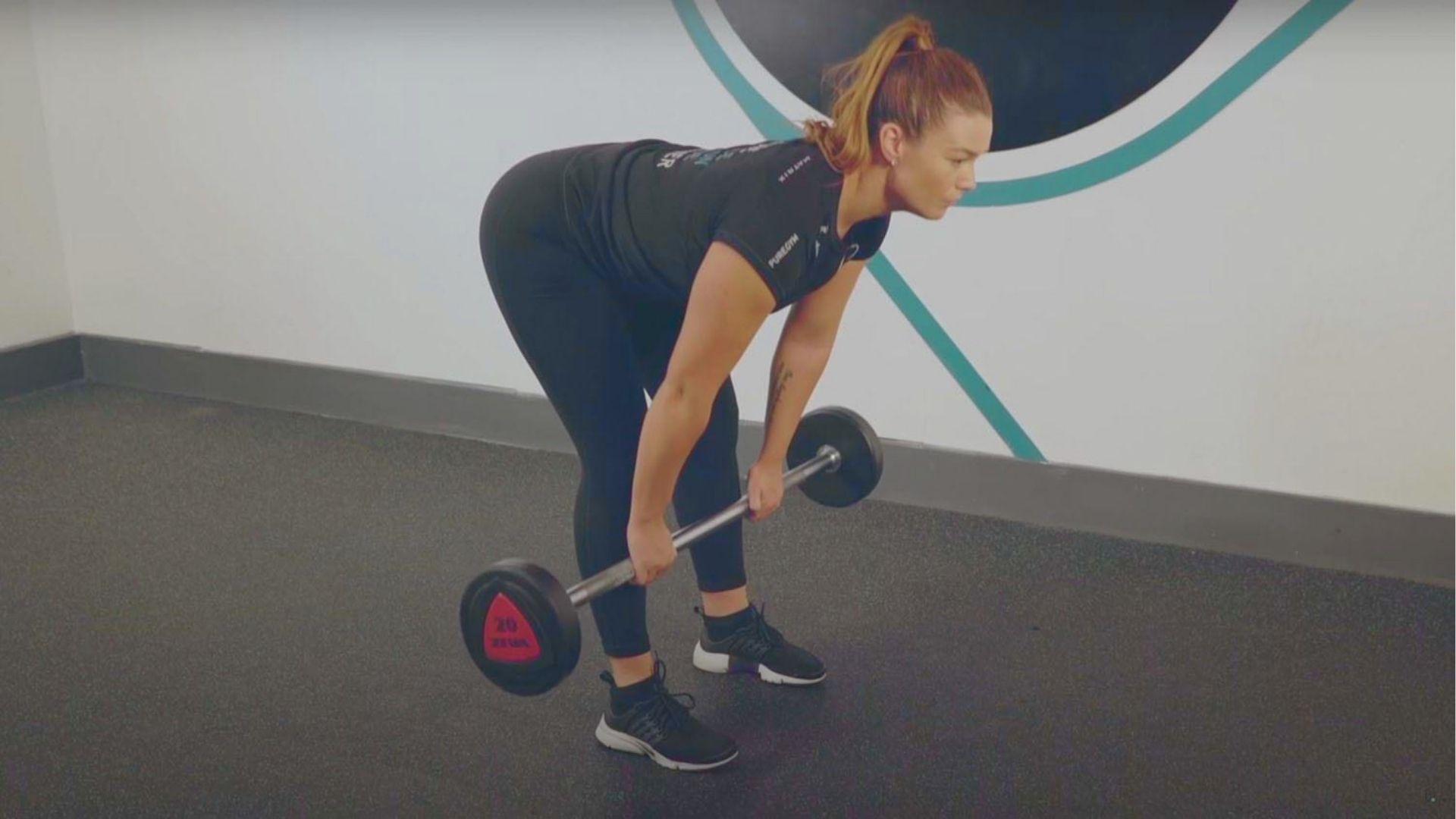 Woman in black PureGym shirt and leggings performing a barbell RDL on gym flooring, lowering weight with hip hinge.